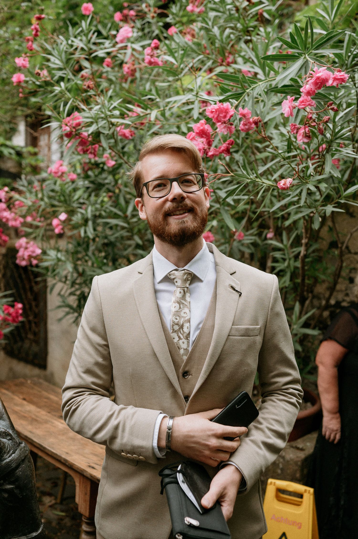 A person in a beige suit stands in front of blooming pink oleander flowers.