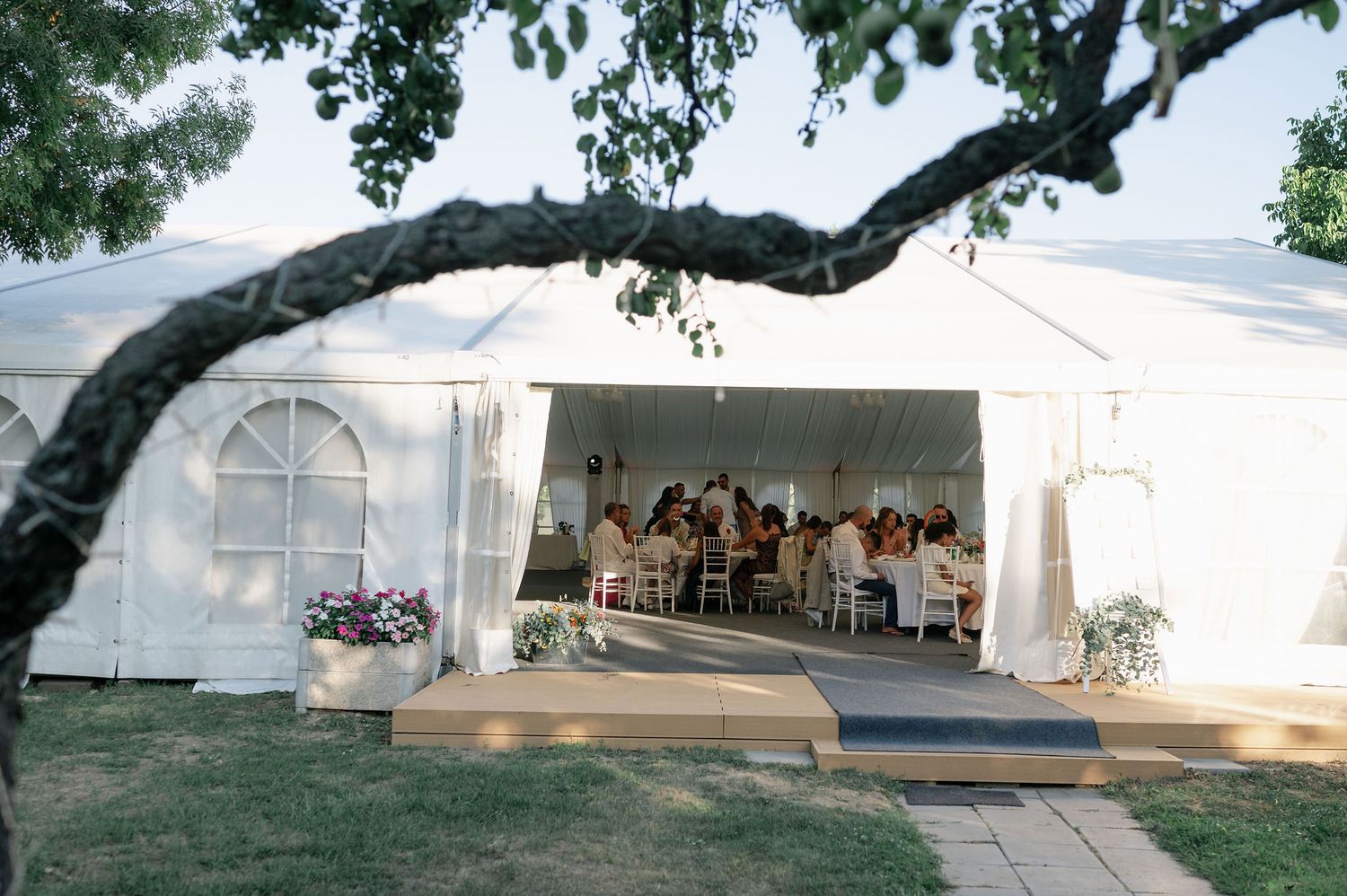 White wedding reception tent set up under large oak trees in a scenic outdoor setting.