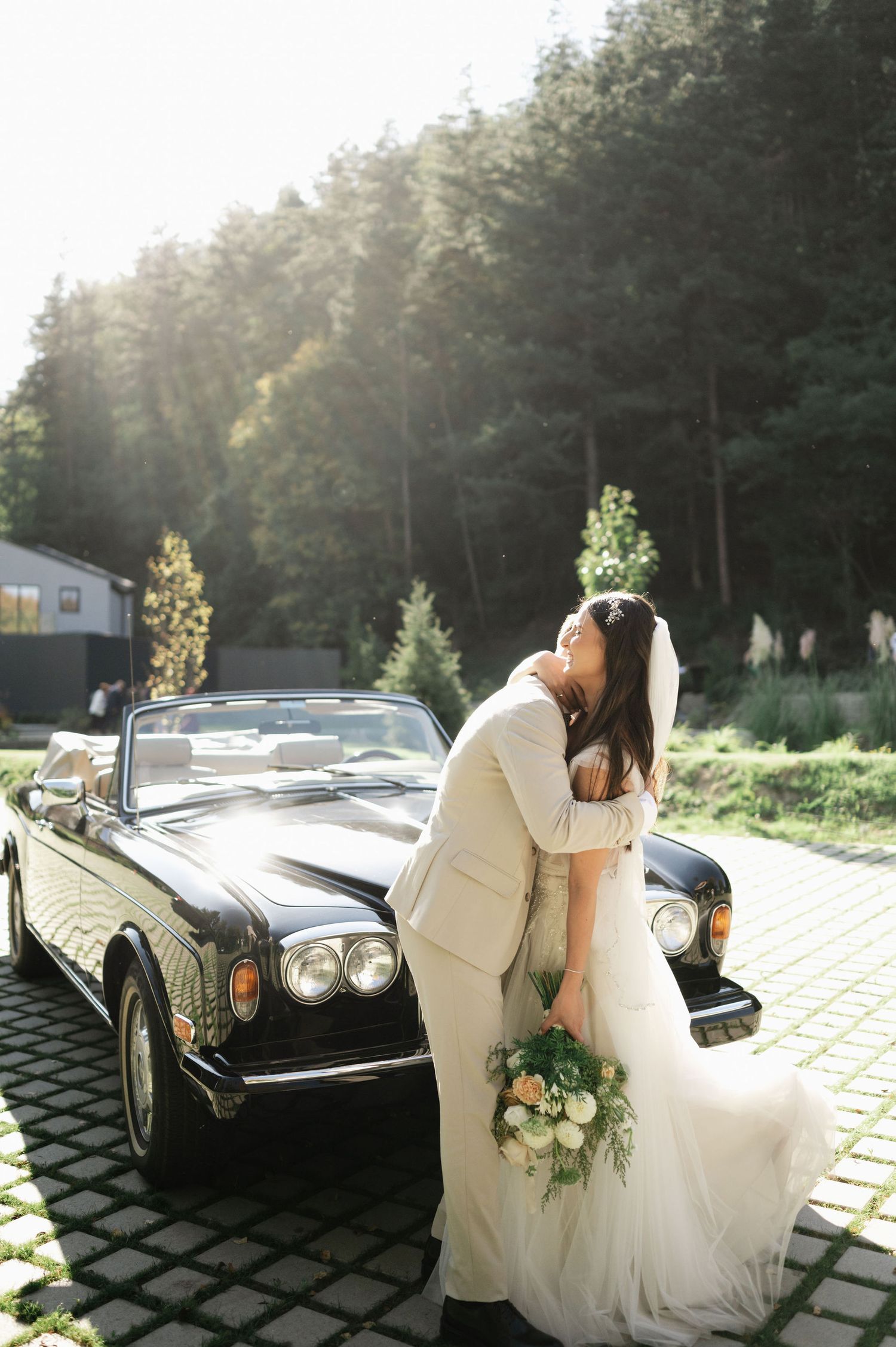 Intimate wedding portrait with classic Rolls Royce against scenic wooded backdrop.