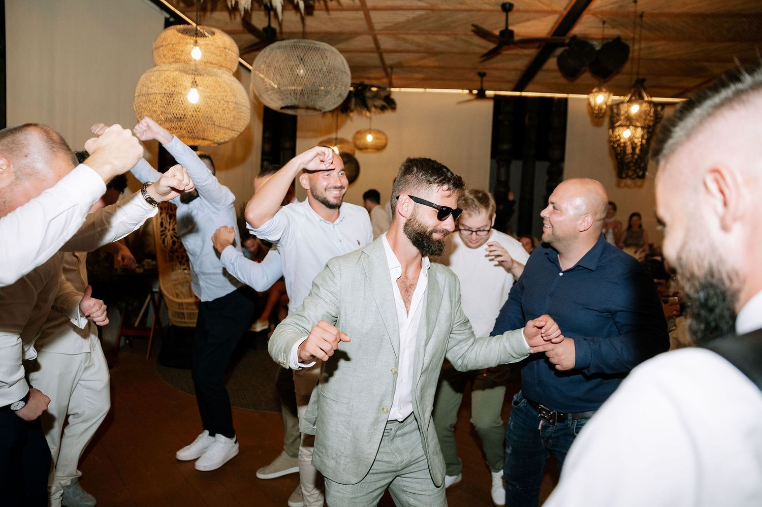 Wedding party members in light-colored suits dancing and celebrating together.