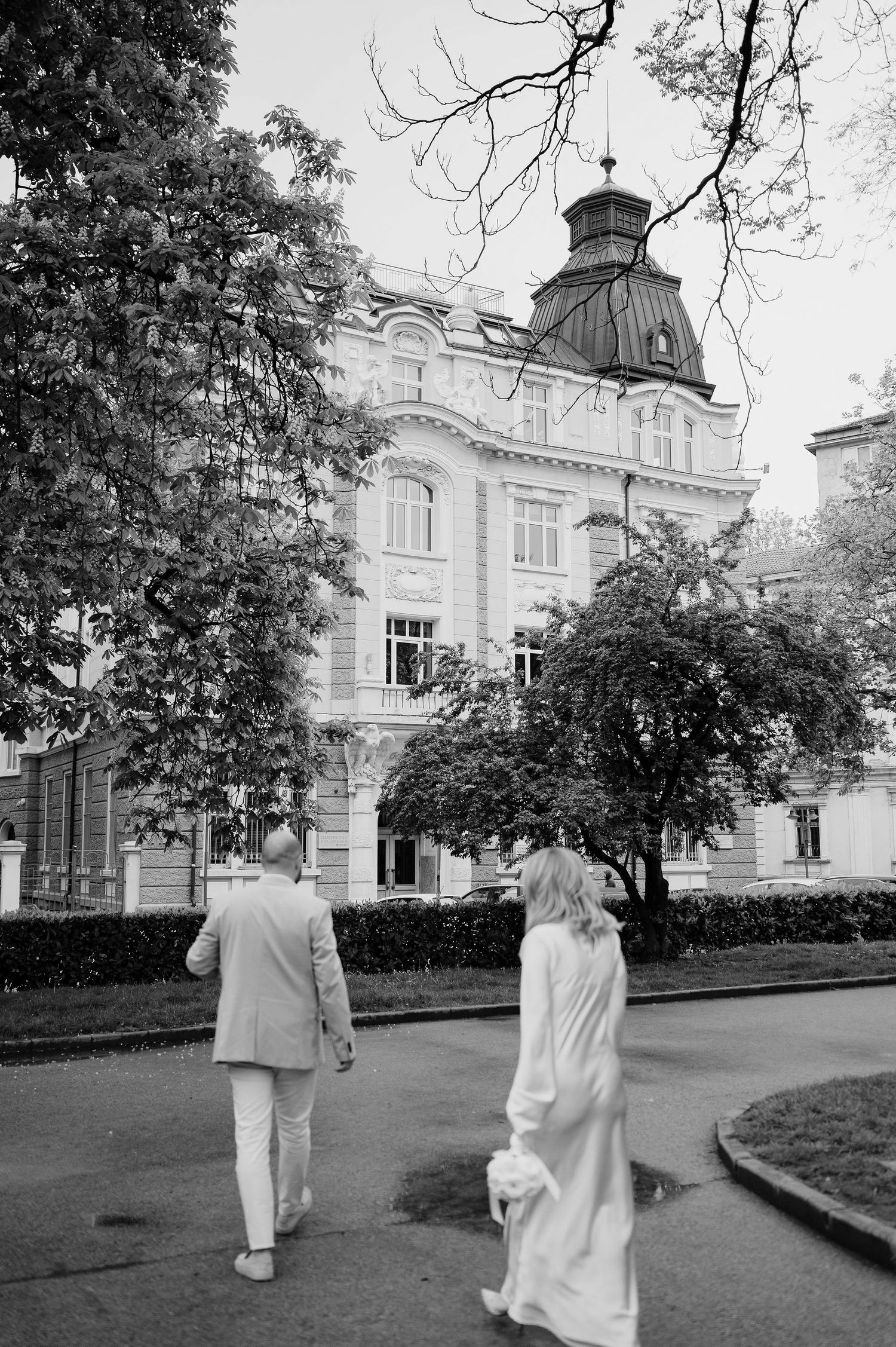 A couple in white clothing walks away from a grand historic building with ornate architecture.