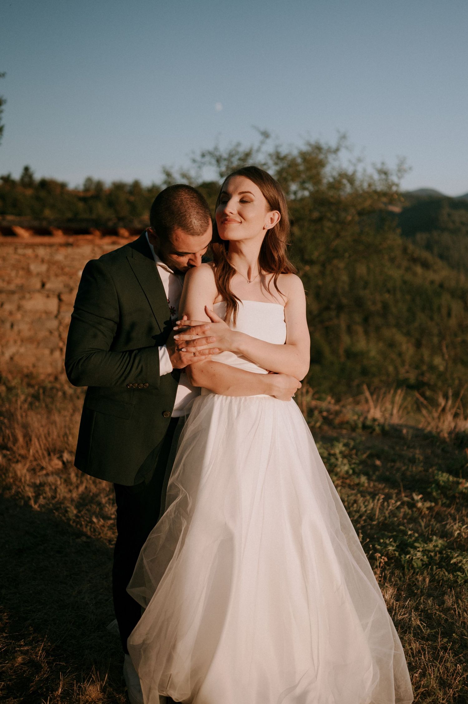A romantic wedding couple embraces against a sunset mountain backdrop.