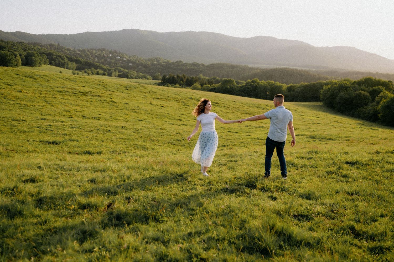 Couple walks through tall grass on a scenic mountainside at sunset.