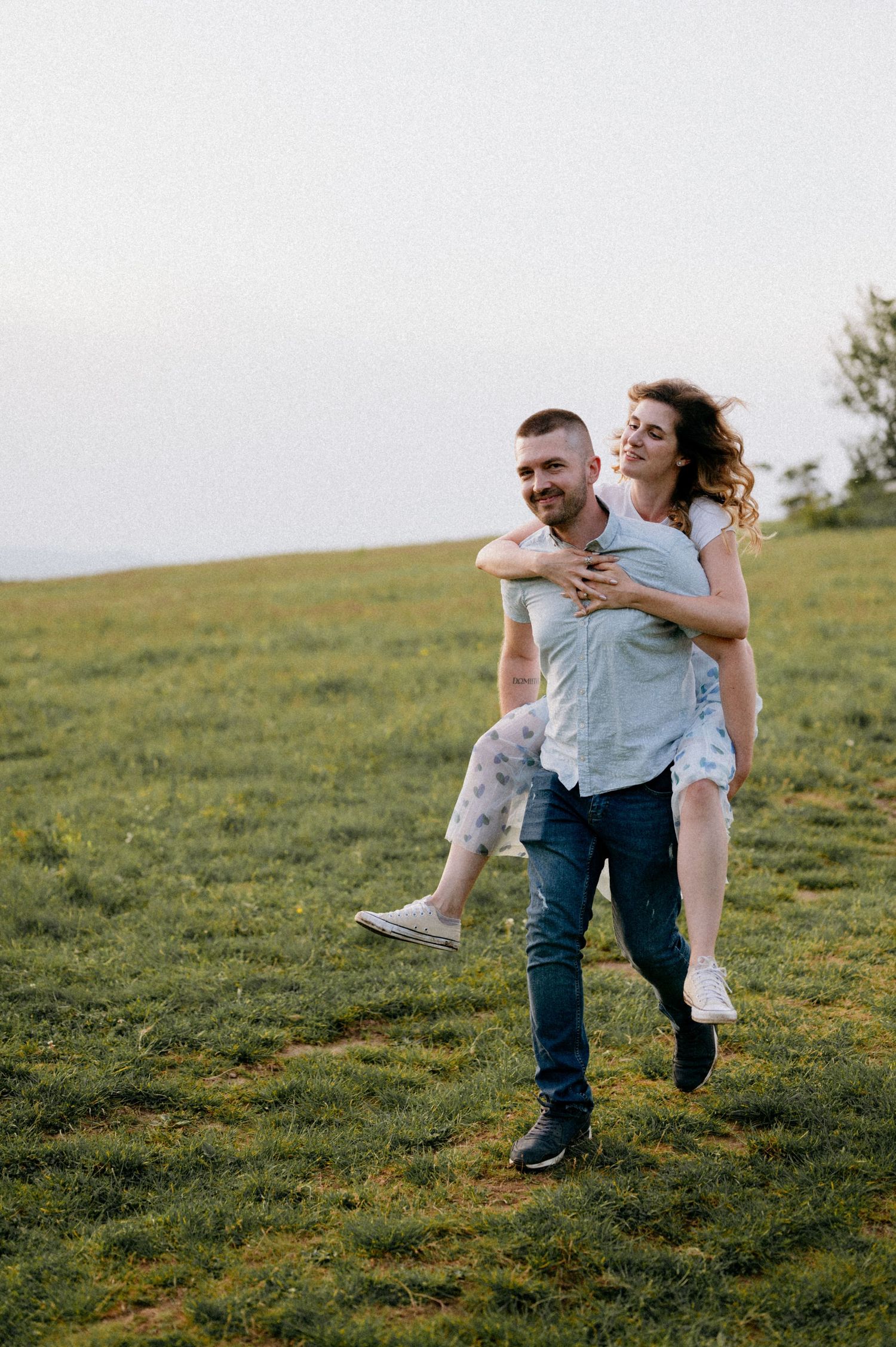 A couple shares a playful piggyback ride moment in a scenic meadow.