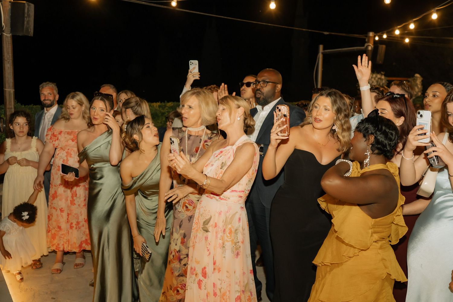 Guests in formal attire celebrate at an outdoor wedding reception at night, holding phones and dancing under string lights.