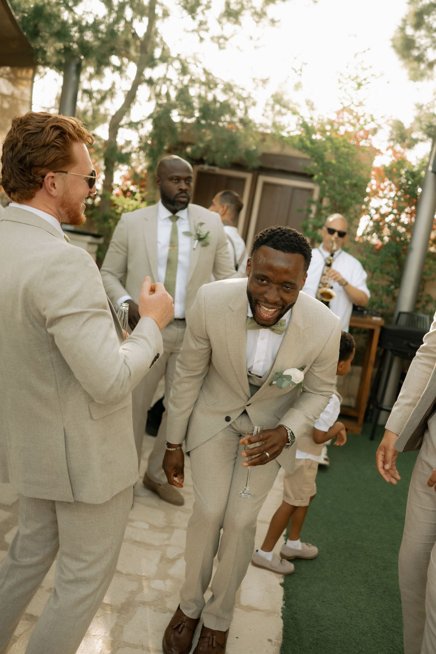 Groomsmen in matching beige suits laughing and celebrating outdoors at a wedding reception.