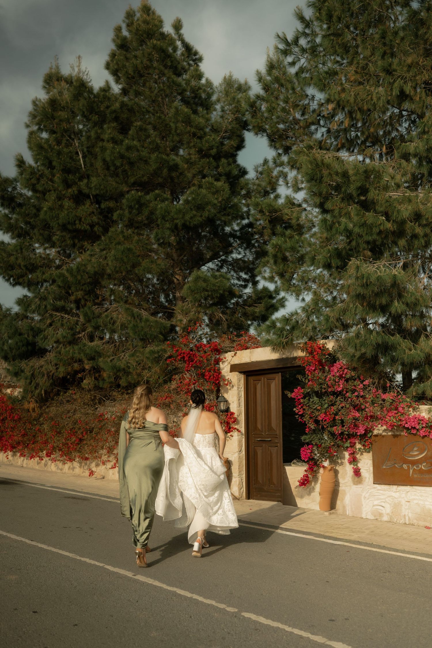 Bride in white gown and bridesmaid in green dress walking toward a Mediterranean venue with bougainvillea.