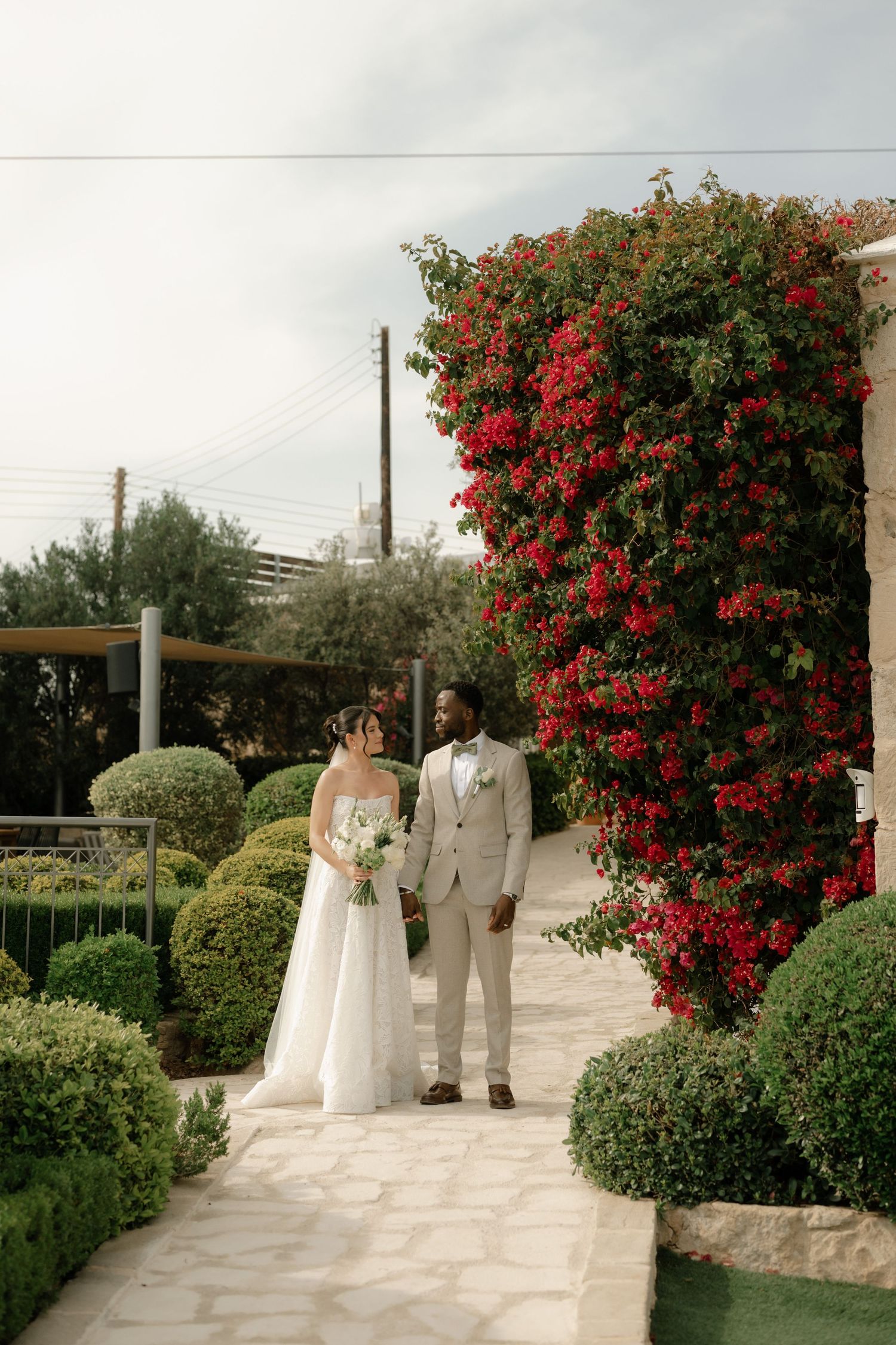 A bride and groom stand together in a lush garden beside vibrant red bougainvillea on their wedding day.