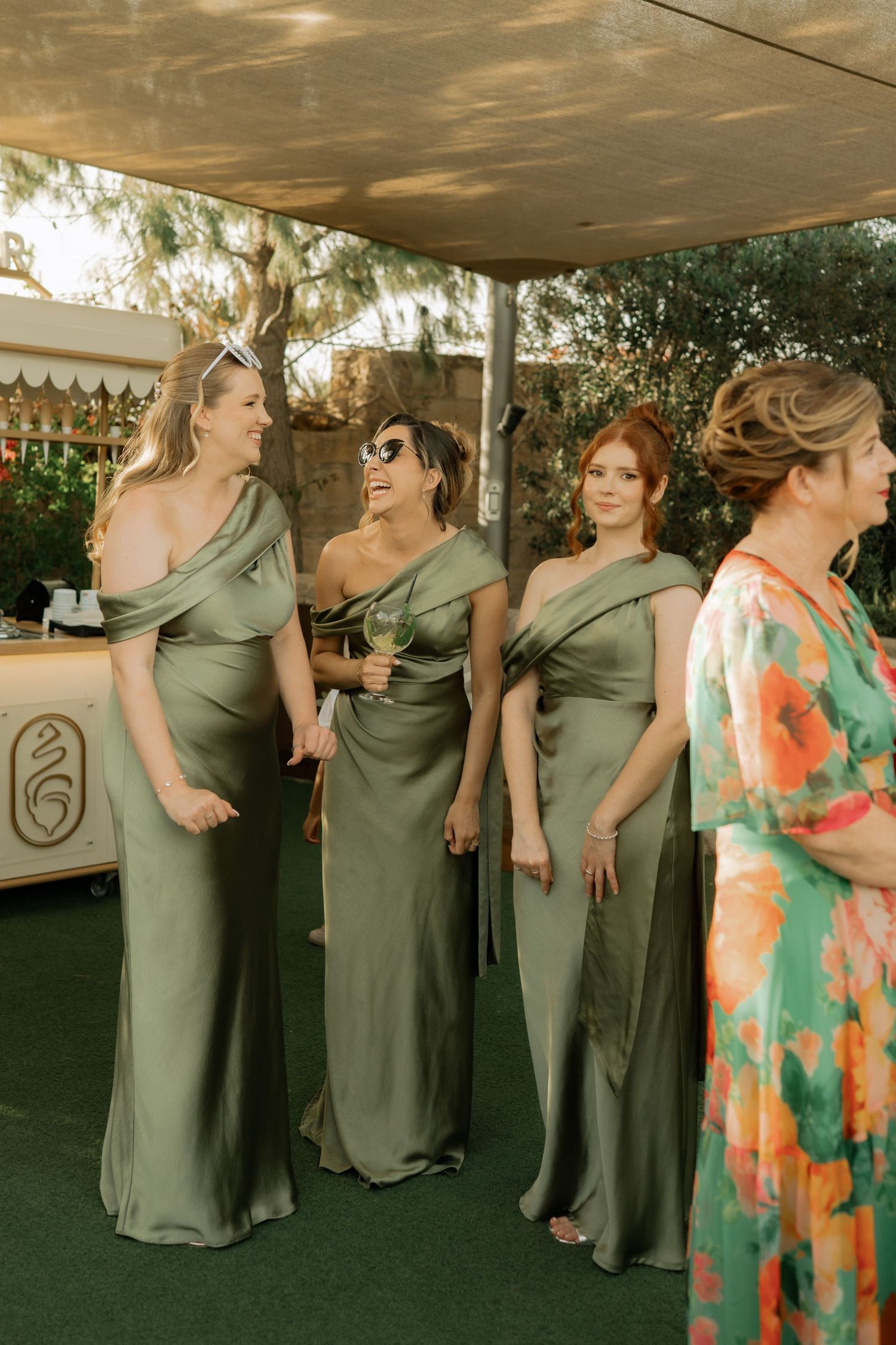 Three bridesmaids in elegant olive green satin gowns standing together at an outdoor wedding reception under a canopy.