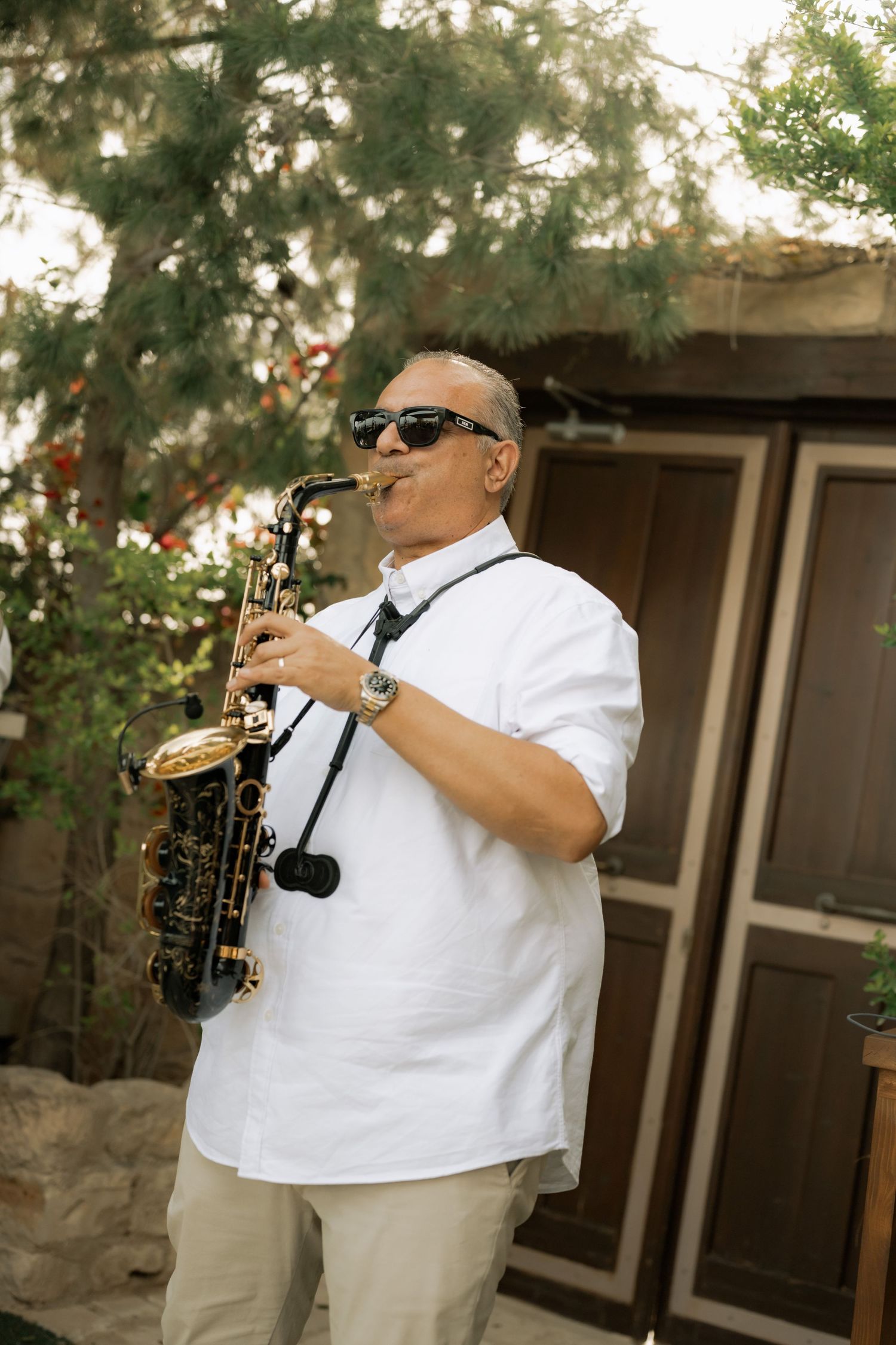 Middle-aged man in white shirt and sunglasses playing a black and gold saxophone outdoors near green trees.