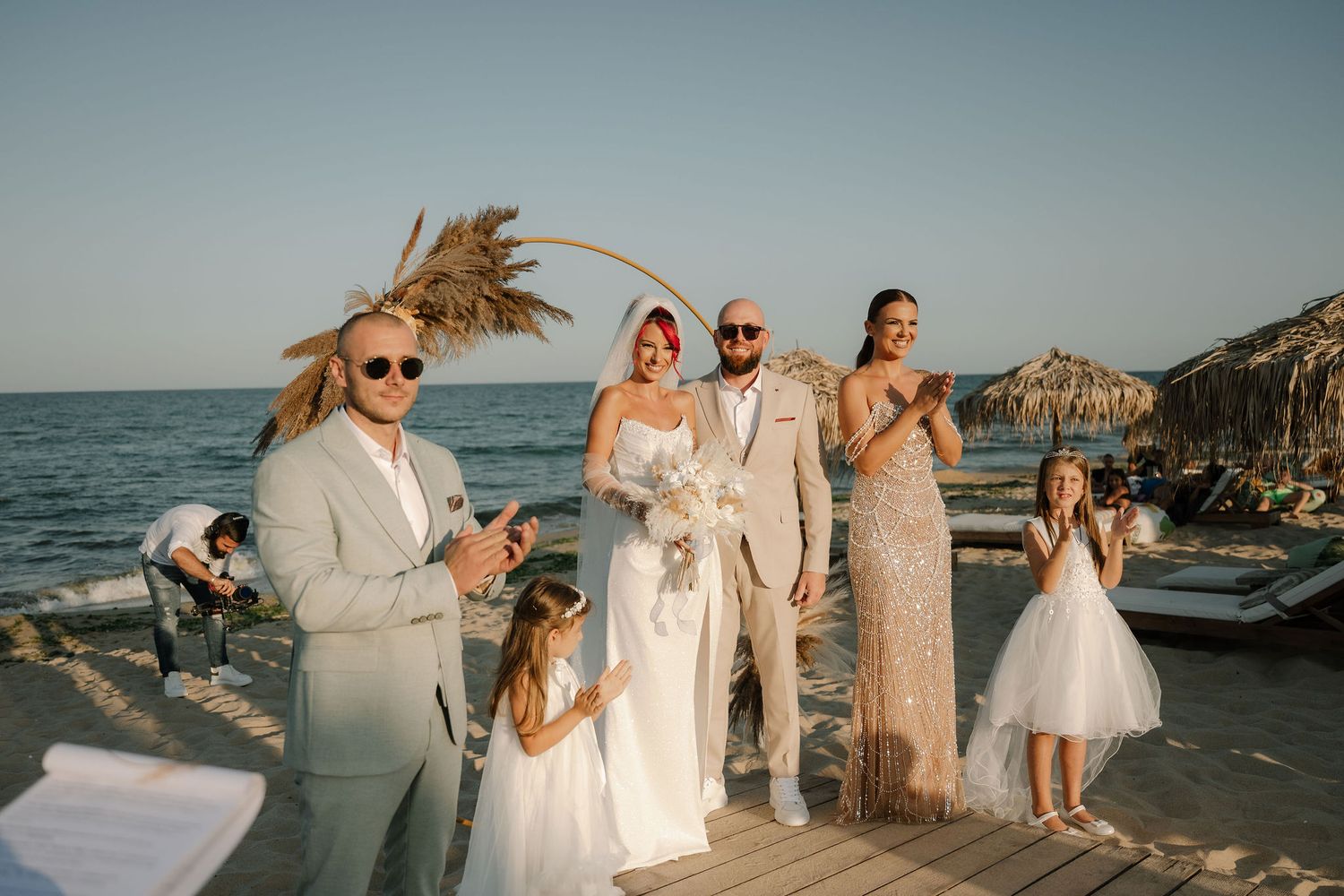 A beachside wedding celebration with guests gathered on a coastal deck at sunset with palm trees and ocean views.