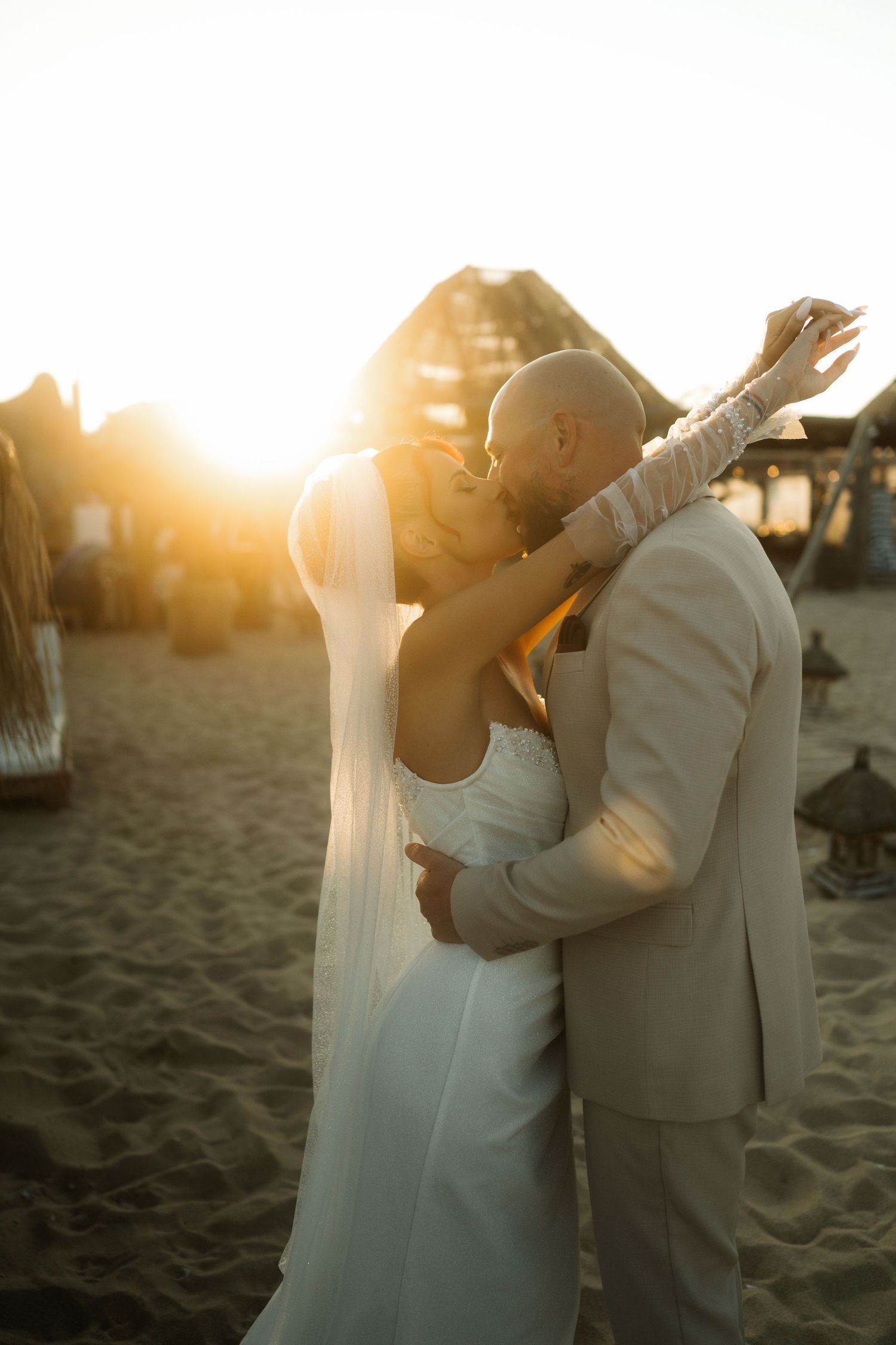 A couple embraces in a tender moment on the sandy beach as the warm sunset light glows behind them.
