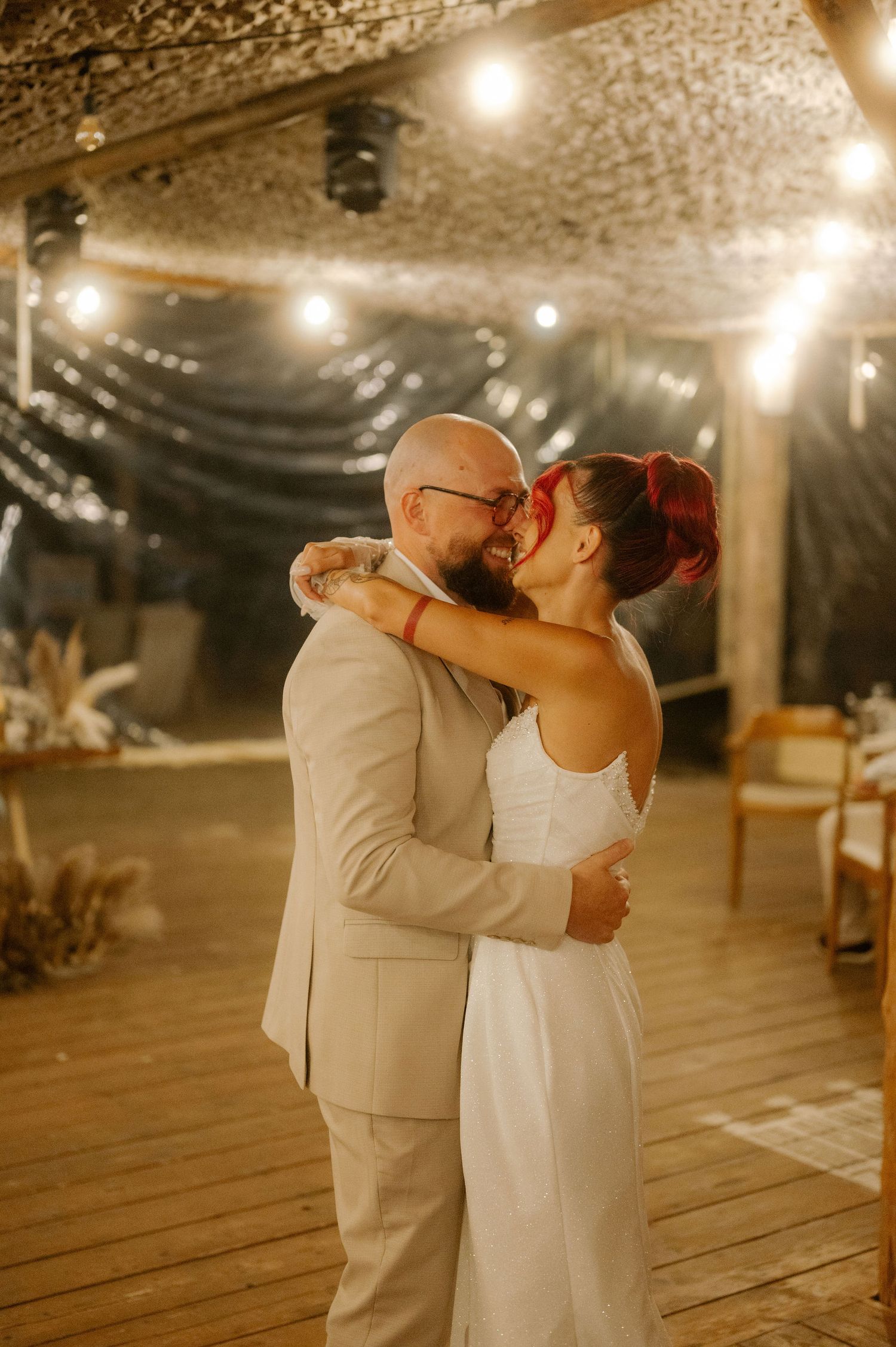 A couple shares their first dance together at a rustic indoor wedding reception with warm lighting and wooden floors.