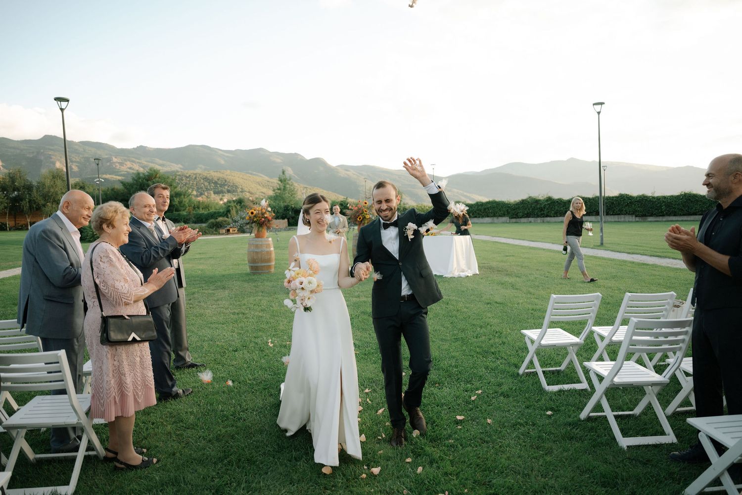 Wedding guests gather on a grassy lawn with white chairs during an outdoor ceremony at sunset in a mountain setting.