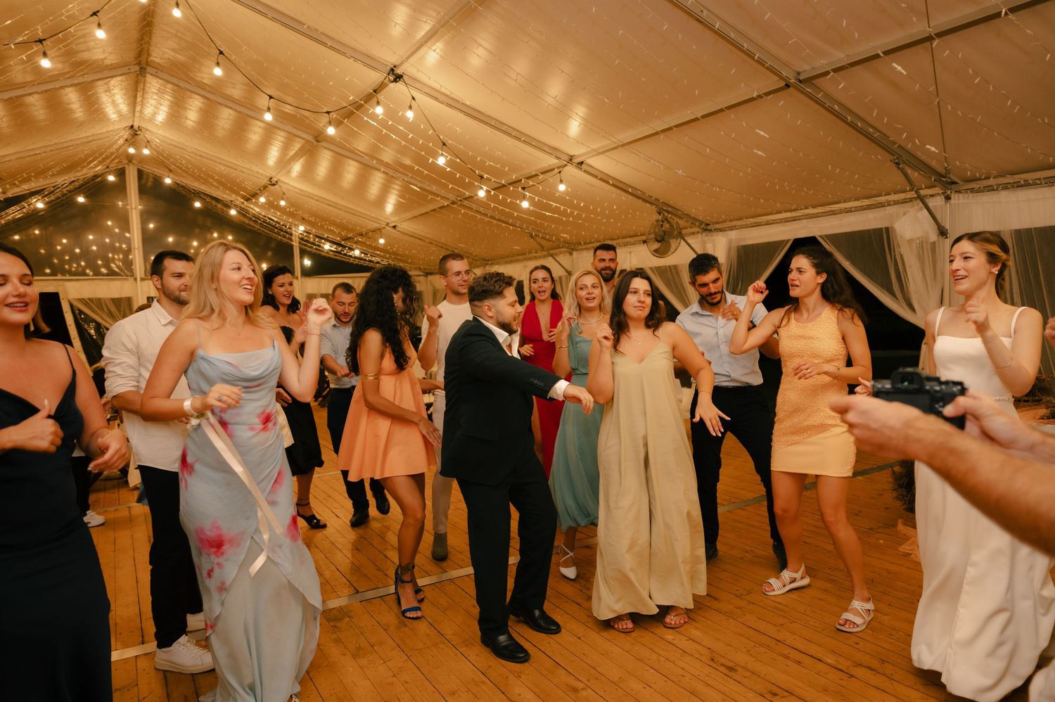 Wedding guests dance and celebrate on a wooden dance floor under warm tent lighting at an evening reception.