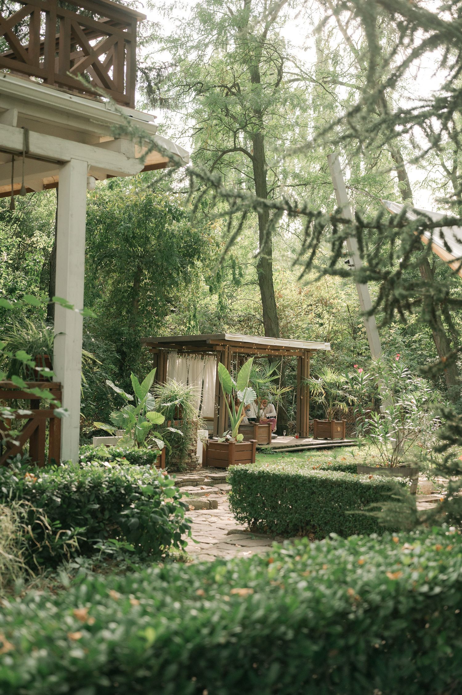 Tranquil garden pathway lined with lush hedges and trees leads to a covered porch area in natural lighting.