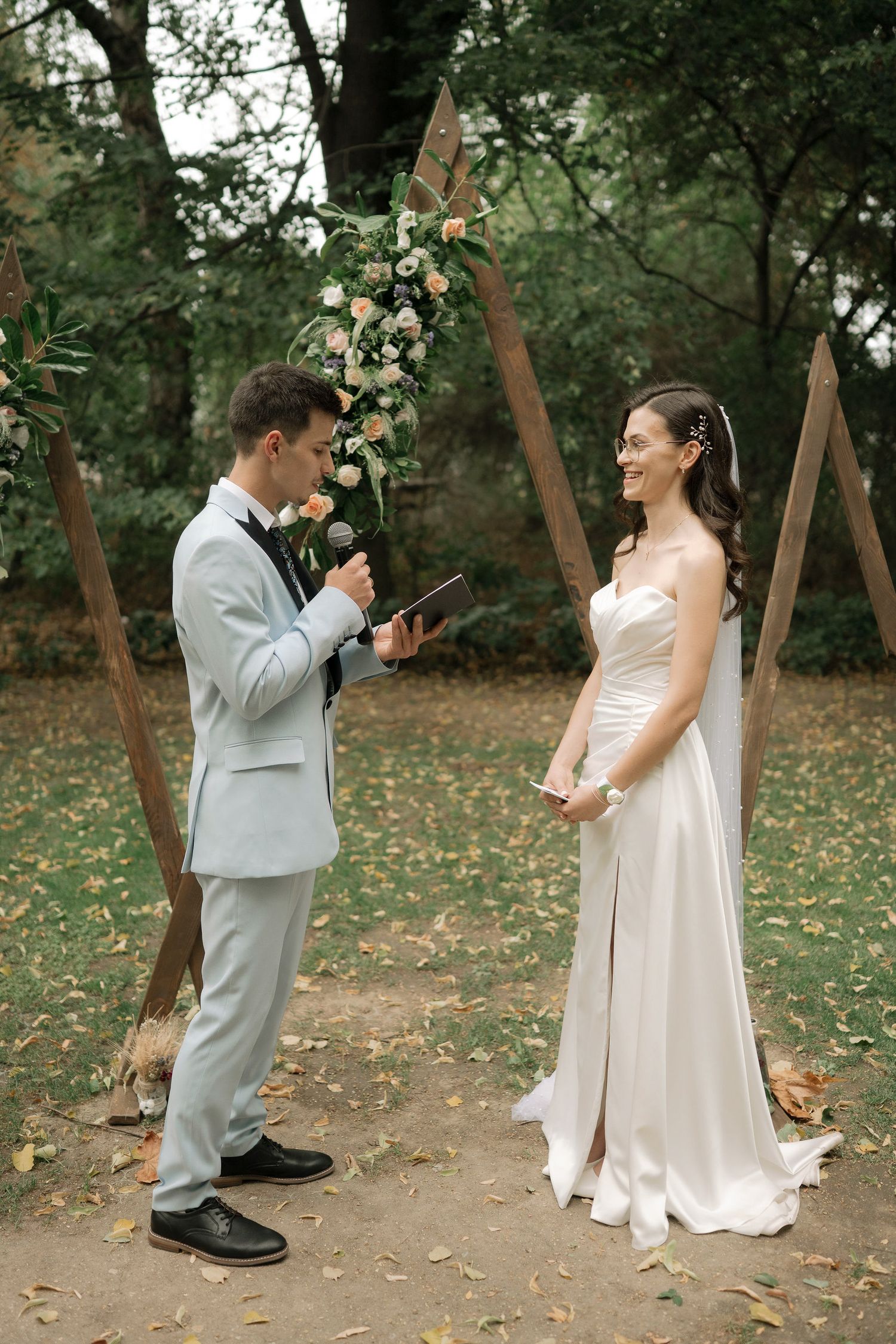 A wedding ceremony takes place outdoors with a wooden triangle arch decorated with white flowers in a wooded setting.