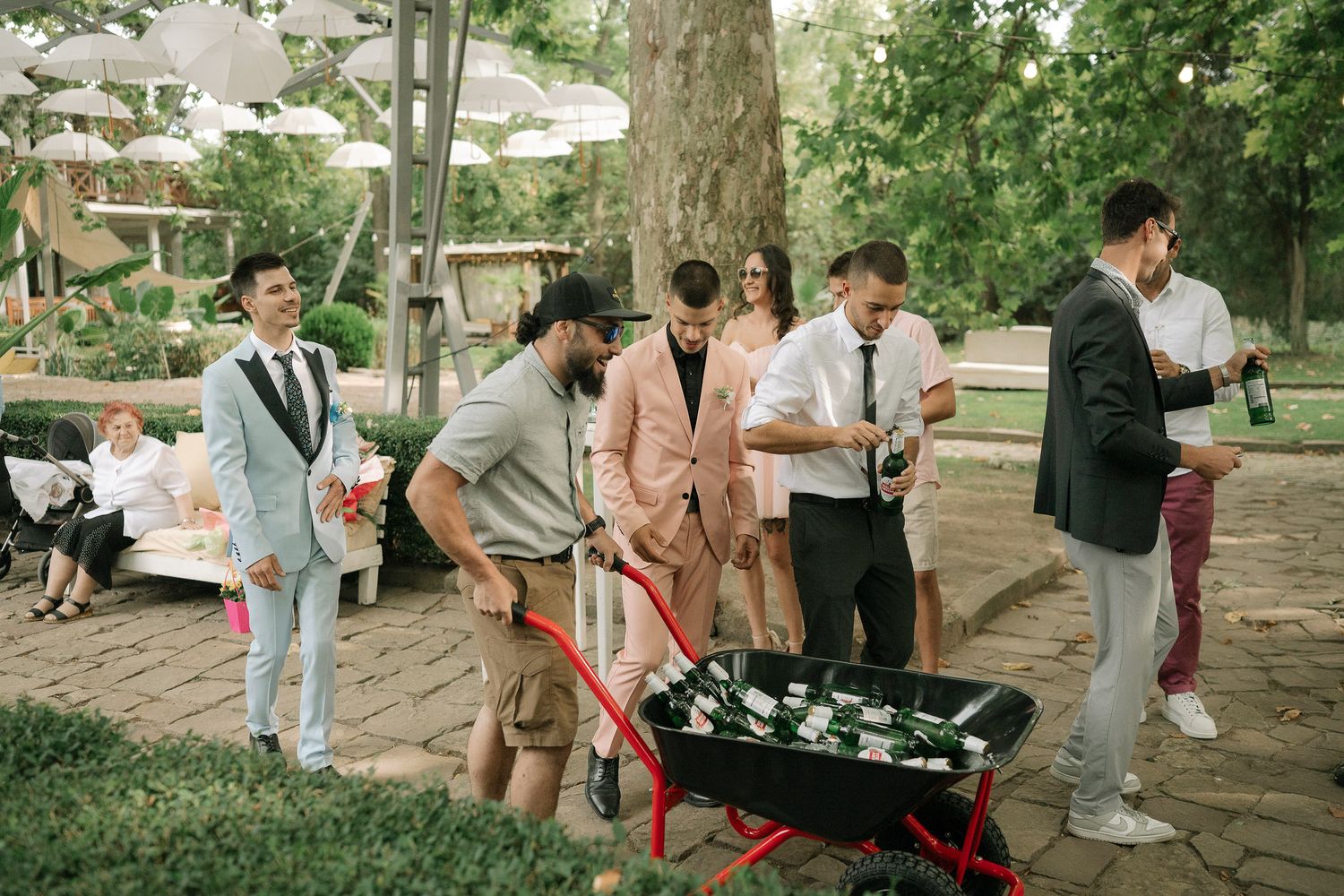 A group gathers around a red wheelbarrow filled with drinks at an outdoor garden party.