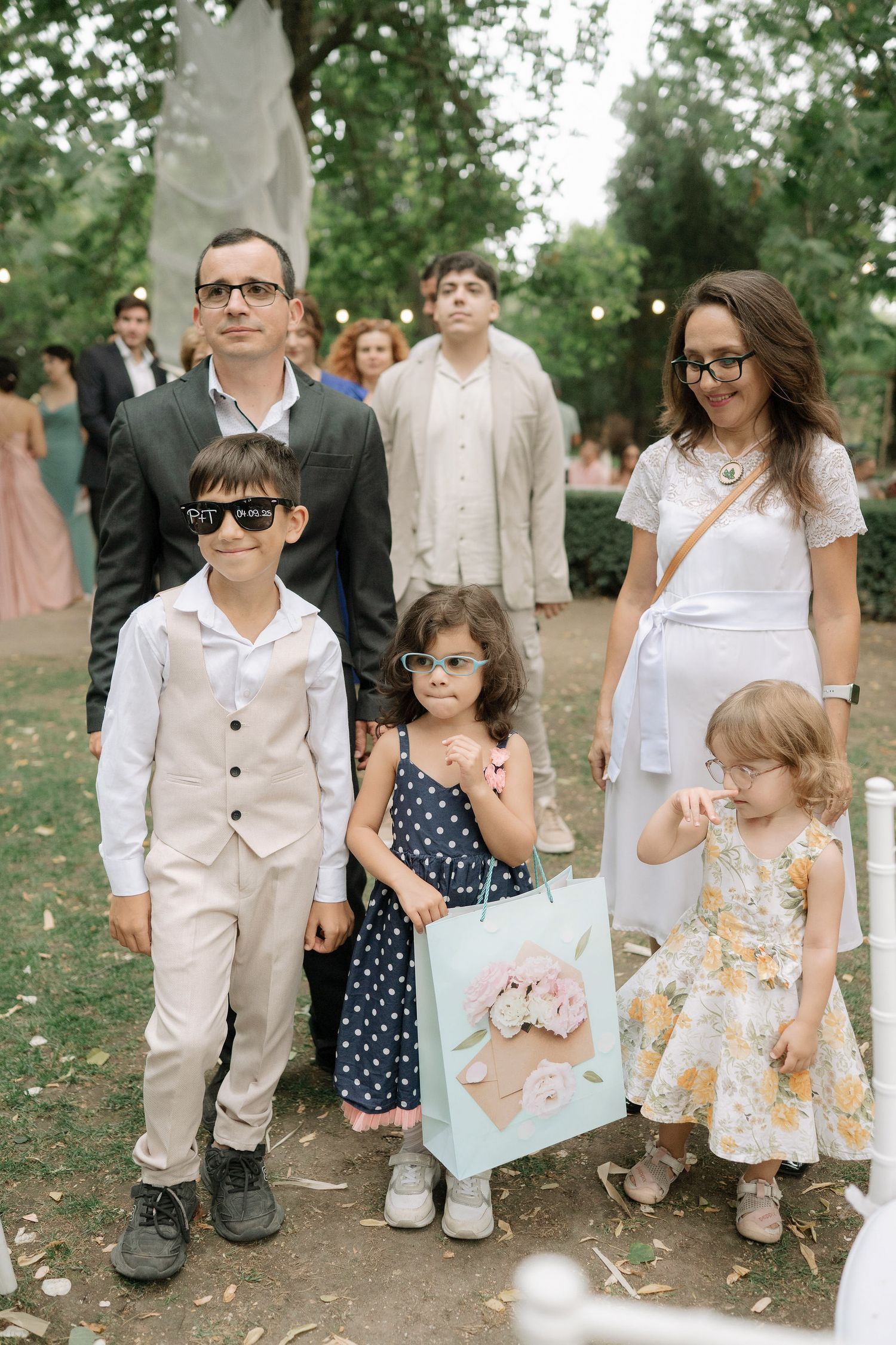 Children in light summer outfits gather outdoors at a formal event in a garden setting.