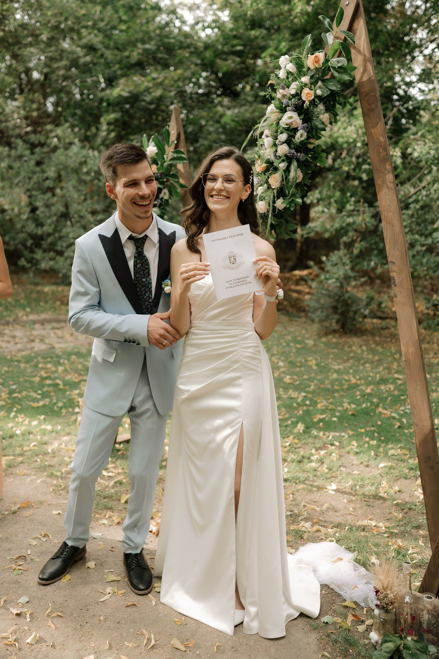 A couple shares joyful moments in their wedding attire under trees in a garden setting.
