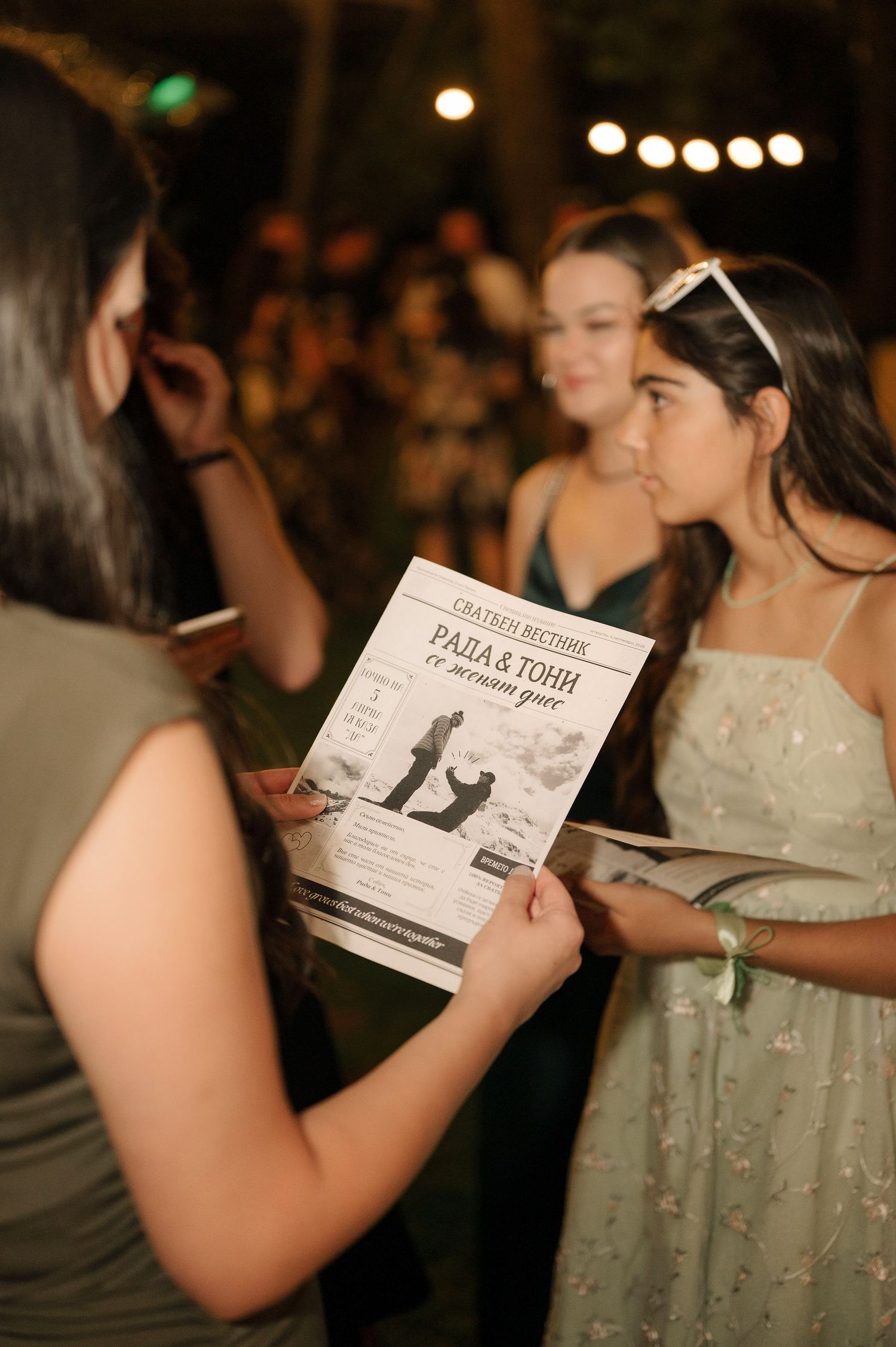 People reading program materials at a nighttime outdoor event with string lights in the background.