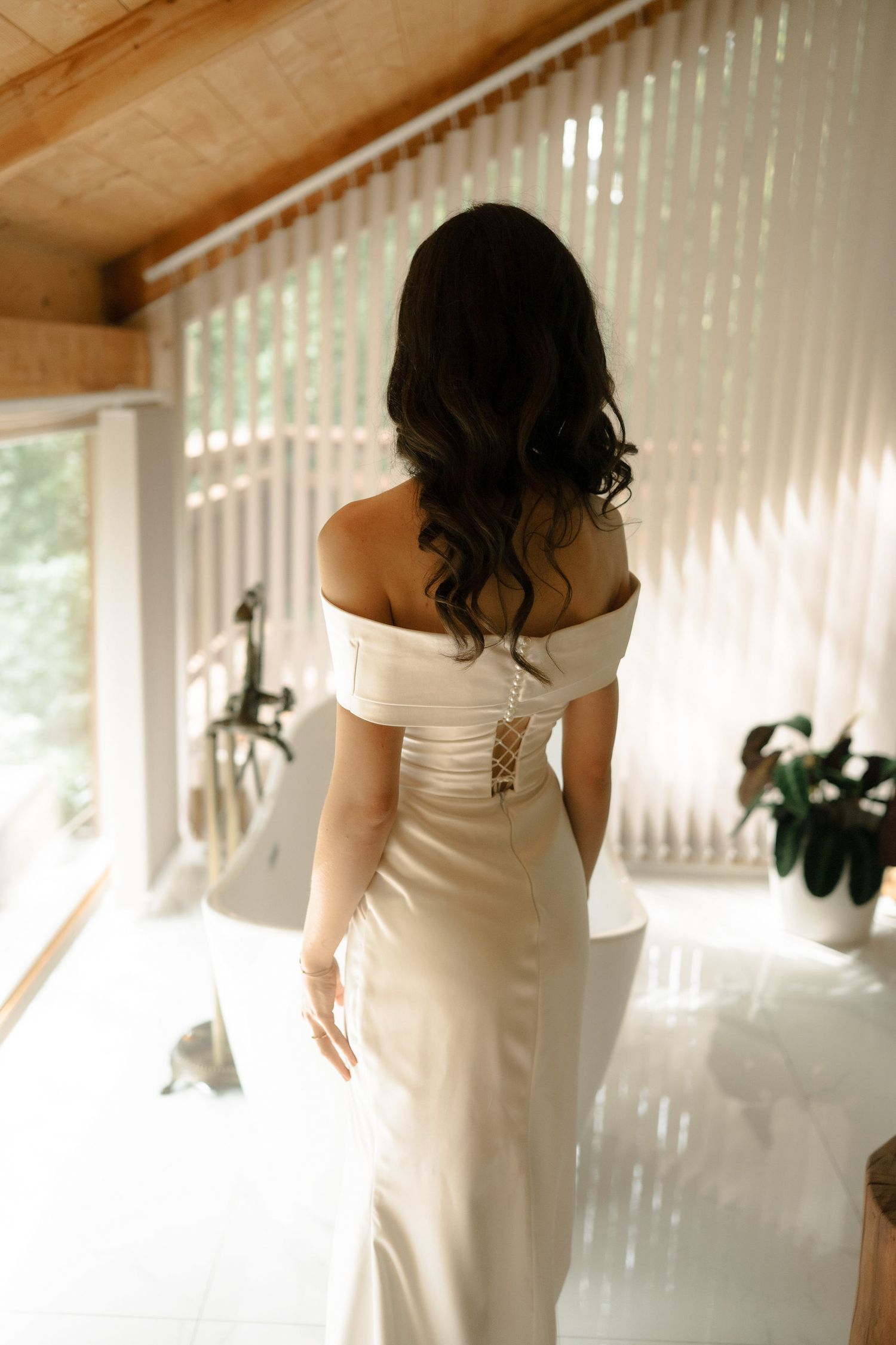 A bride in an elegant white off-shoulder wedding dress stands in soft natural light streaming through vertical blinds.