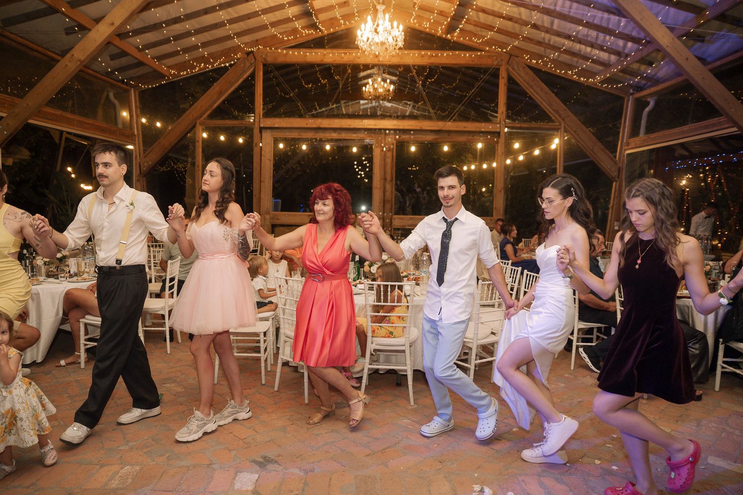 Wedding guests dance in a rustic barn venue decorated with string lights and chandeliers during a reception celebration.