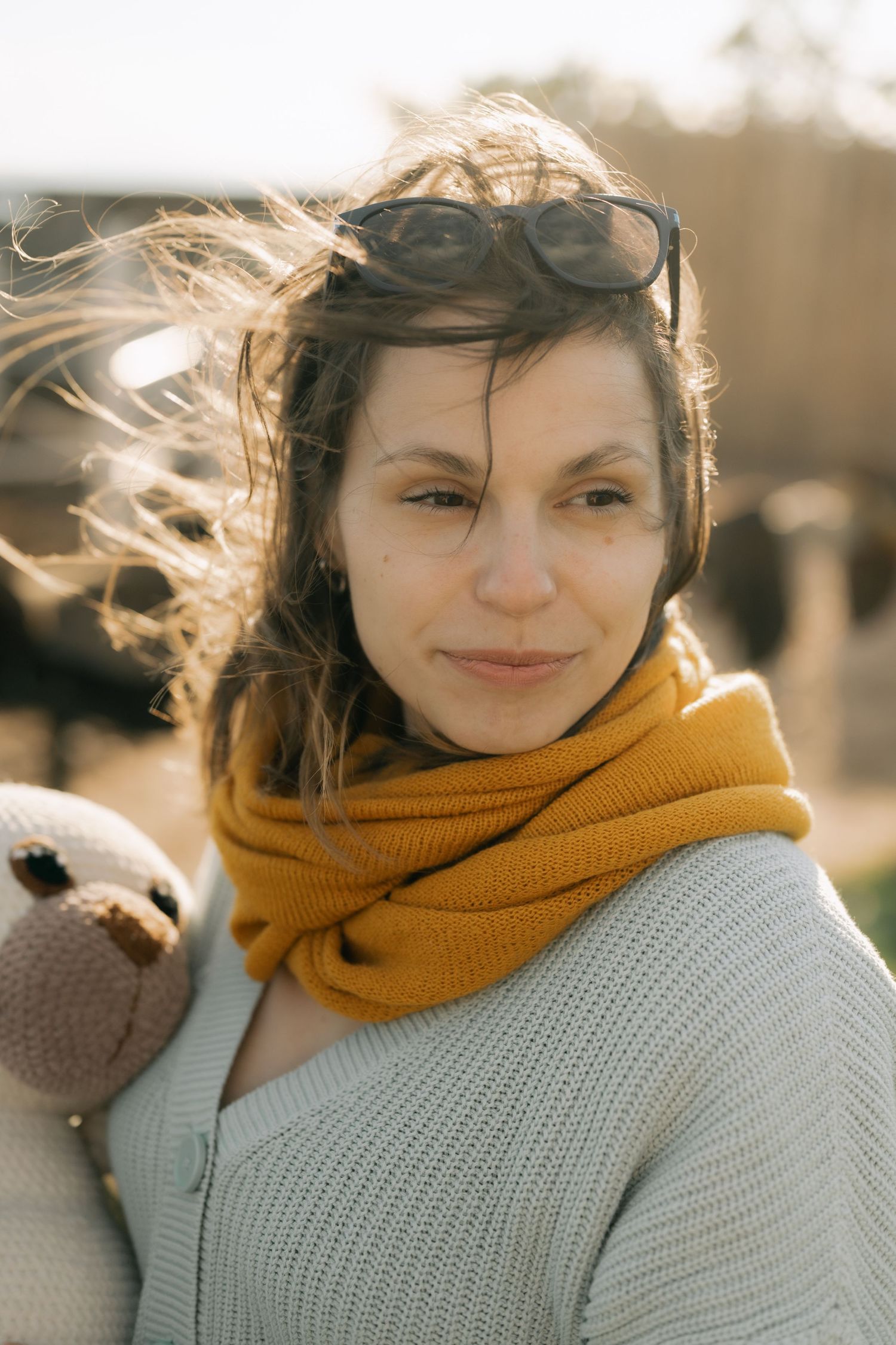 A person wearing a mustard yellow scarf and gray sweater smiles warmly while their hair blows in the wind.