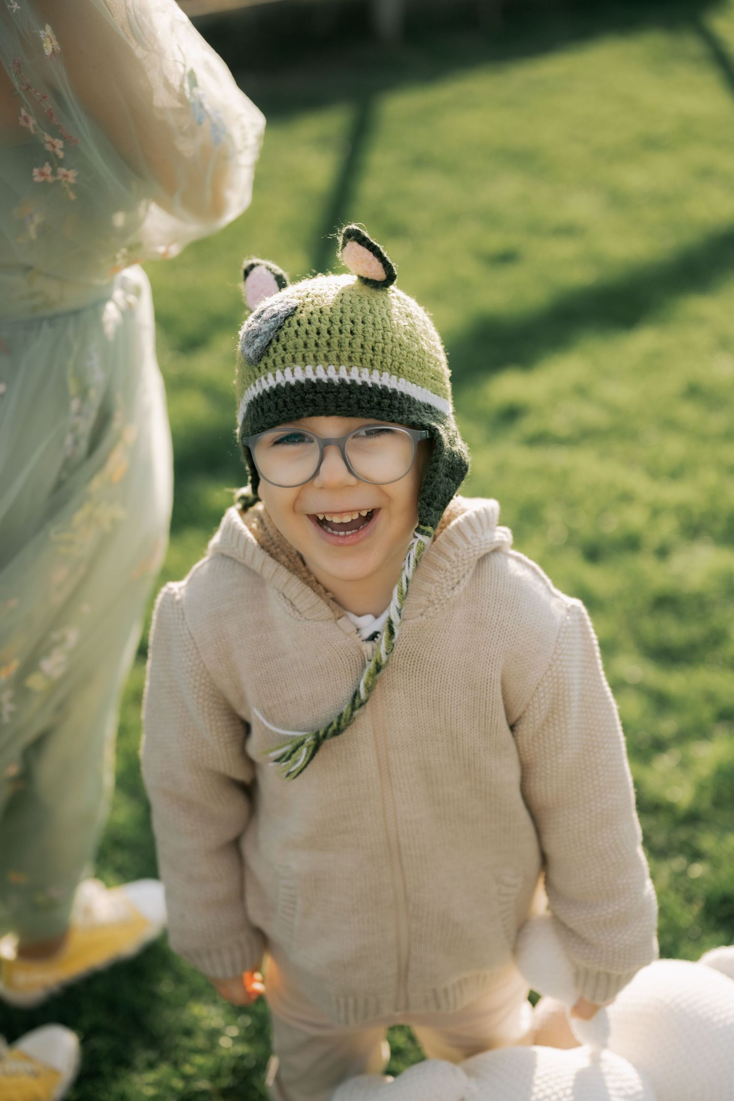 A person wearing a beige sweater and knit hat laughs joyfully while sitting outdoors on a sunny day.