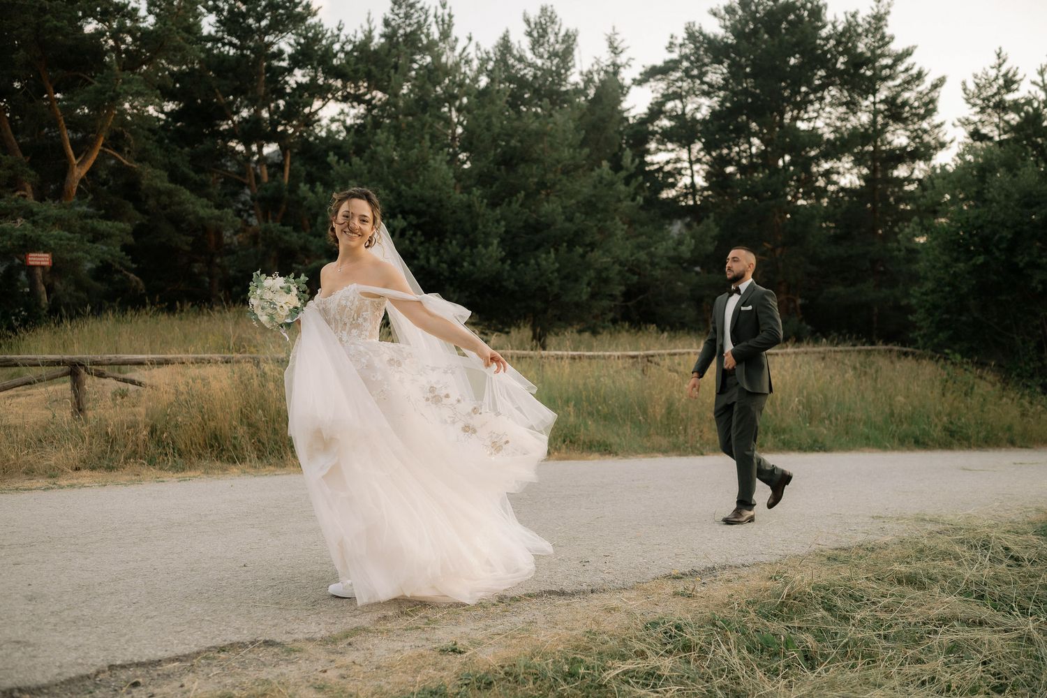 A flowing white wedding dress billows in the wind as figures walk along a mountain path surrounded by evergreens.