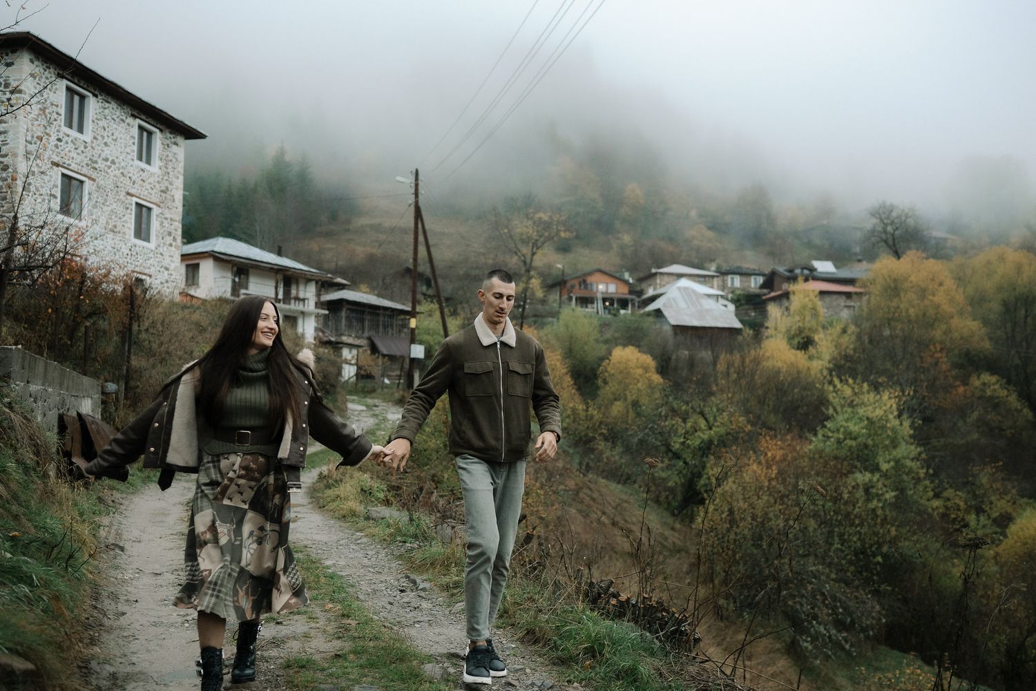 Two people walking on a misty mountain path in autumn with stone buildings and rustic scenery in the background.