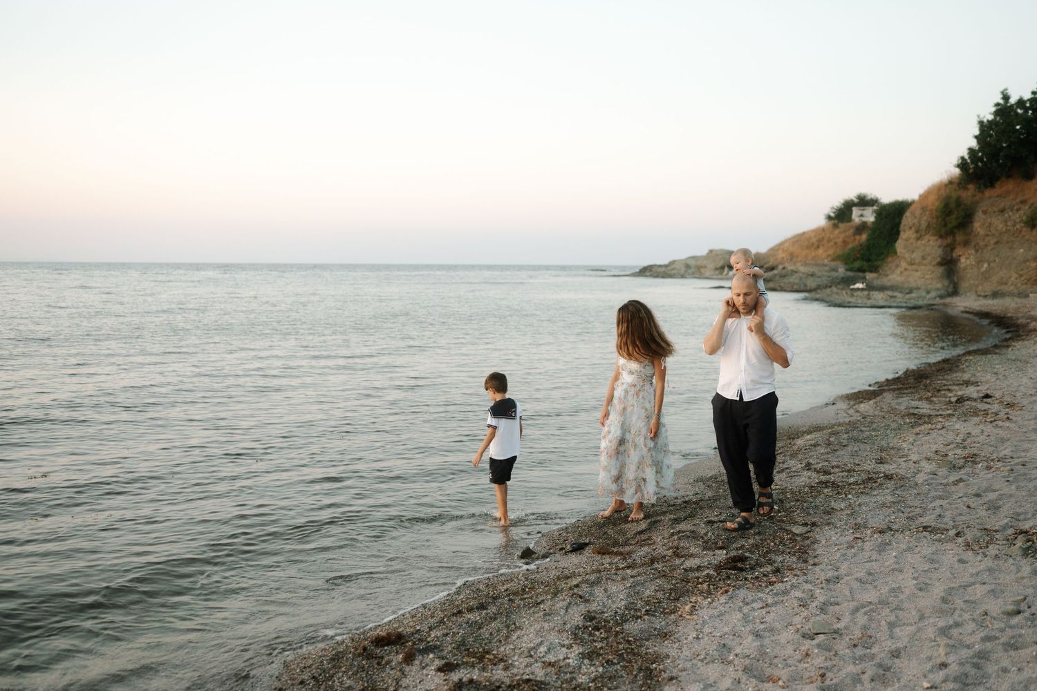 People walking along a scenic coastal beach at sunset with rocky cliffs in the background.