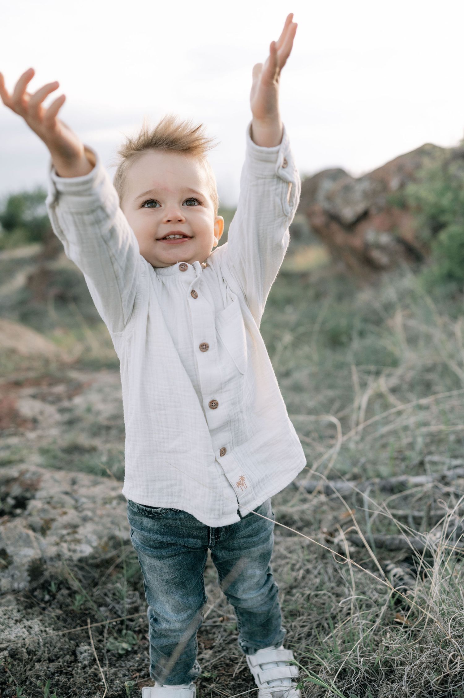 A toddler in a white button-up shirt and jeans plays joyfully outdoors with raised arms in a natural setting.