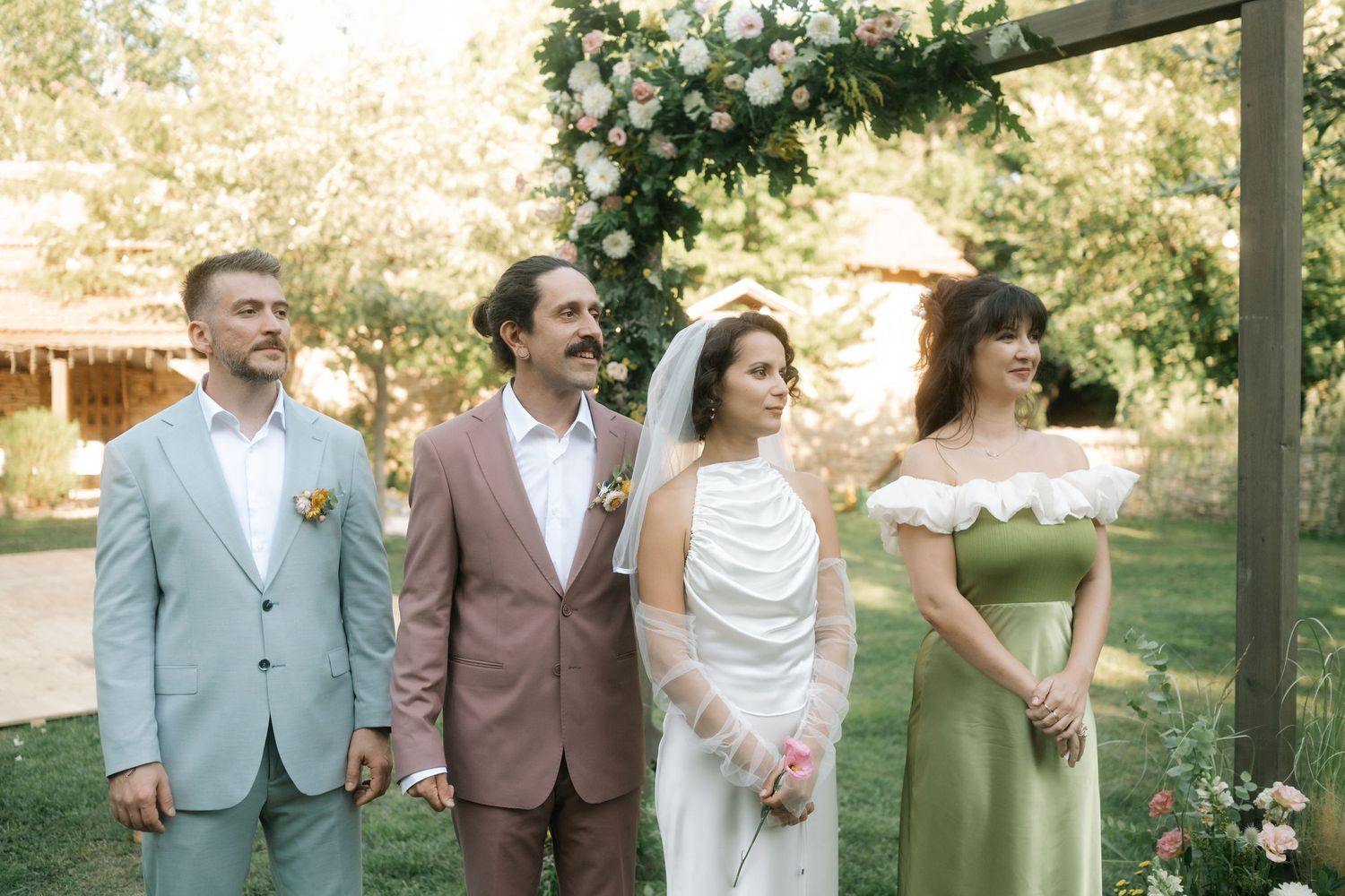 Wedding group poses outdoors under floral-decorated arch with greenery and rustic backdrop.