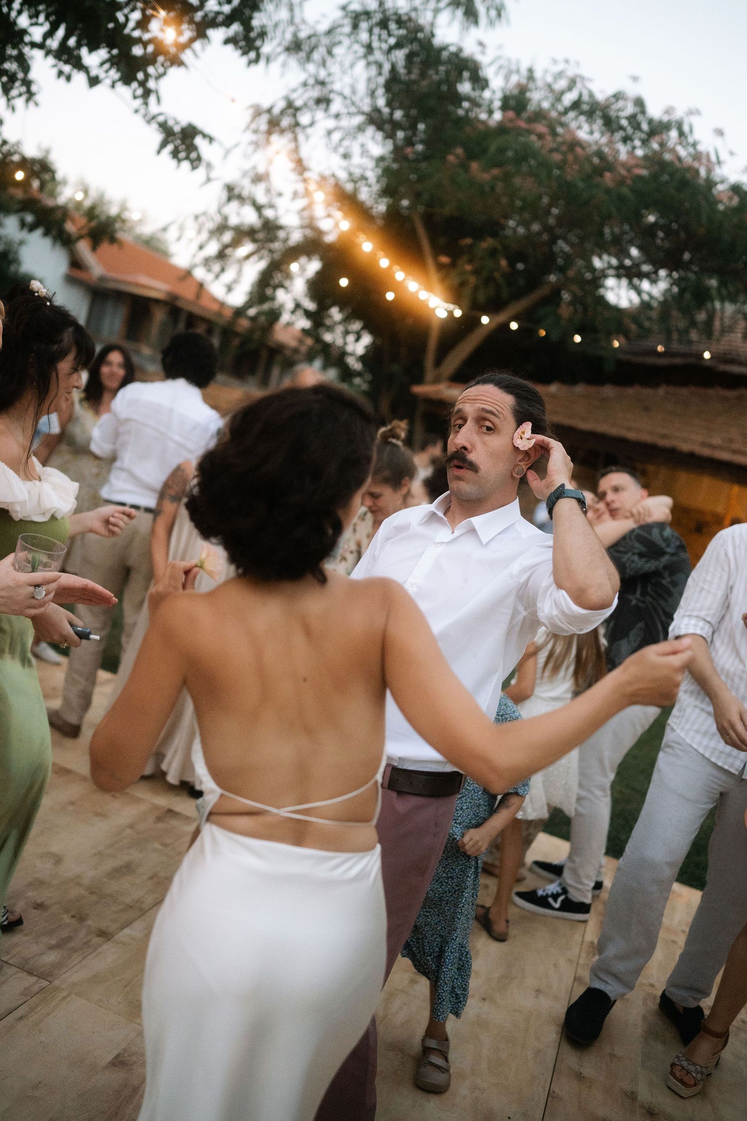 People in white attire dancing and celebrating at an outdoor party with string lights overhead at dusk.