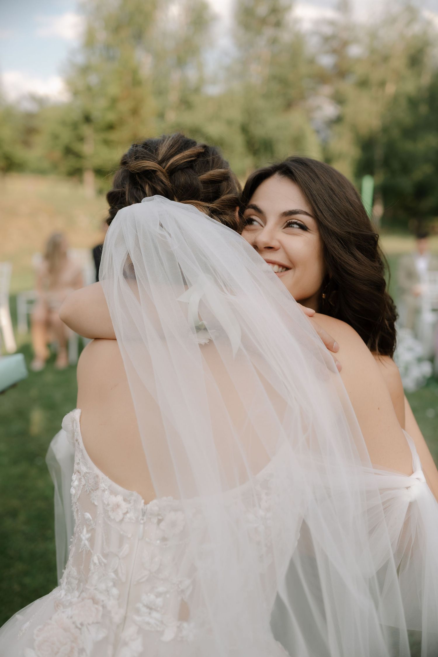 Two people share a joyful embrace at an outdoor wedding ceremony with a flowing white veil and lace dress visible.