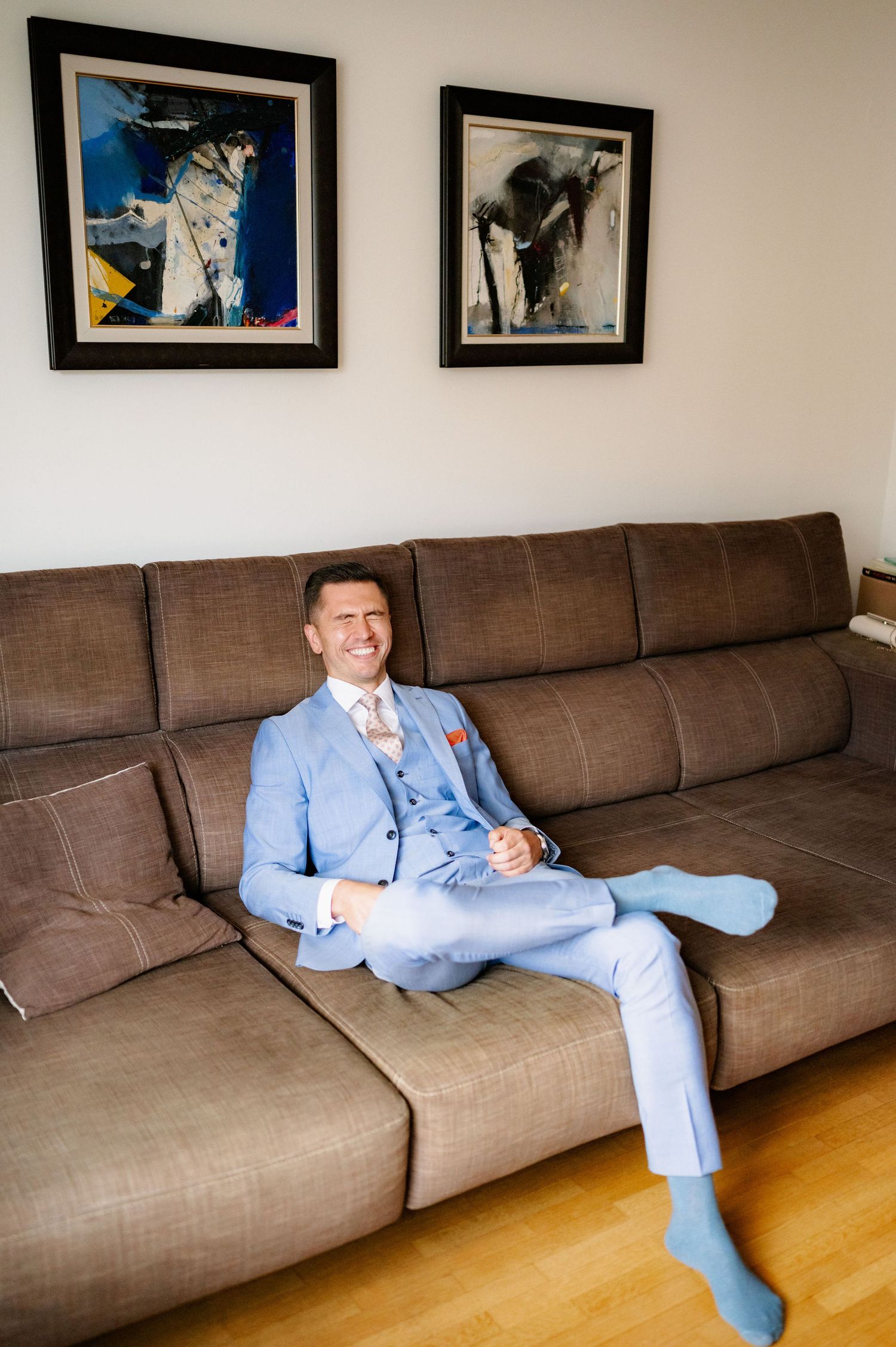 A person in a light blue suit relaxes on a long brown sectional couch with framed artwork on the wall.