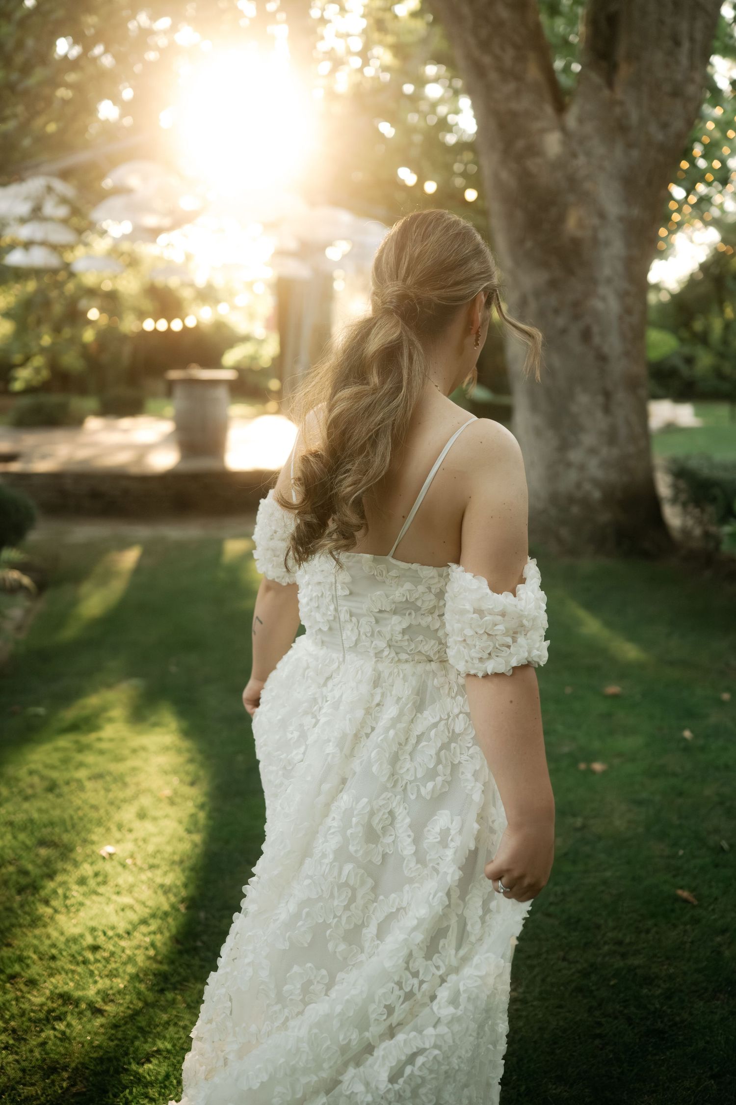 A bride in a textured off-shoulder white wedding gown stands in a sunlit garden during golden hour.