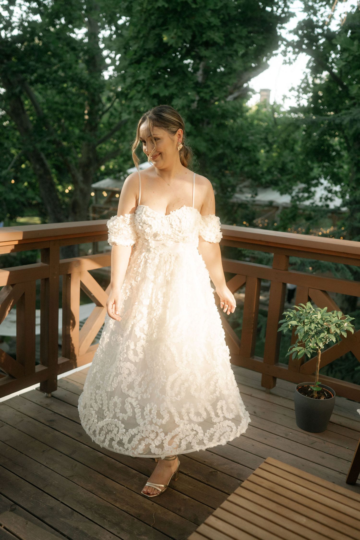 A white lace dress twirls on a wooden deck surrounded by lush green trees and outdoor decor.