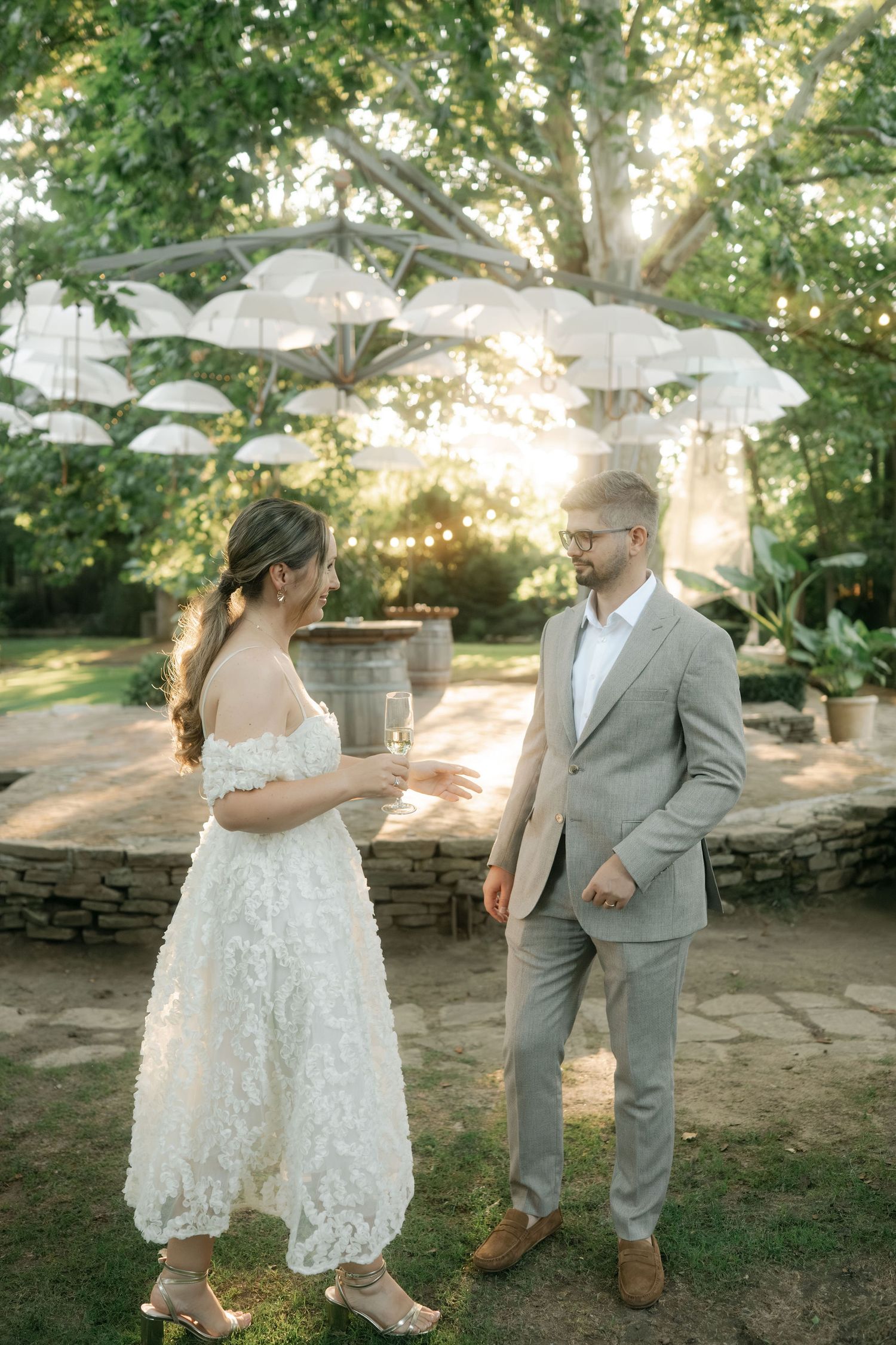 A couple in wedding attire dances together outdoors in a garden setting with natural sunlight filtering through trees.