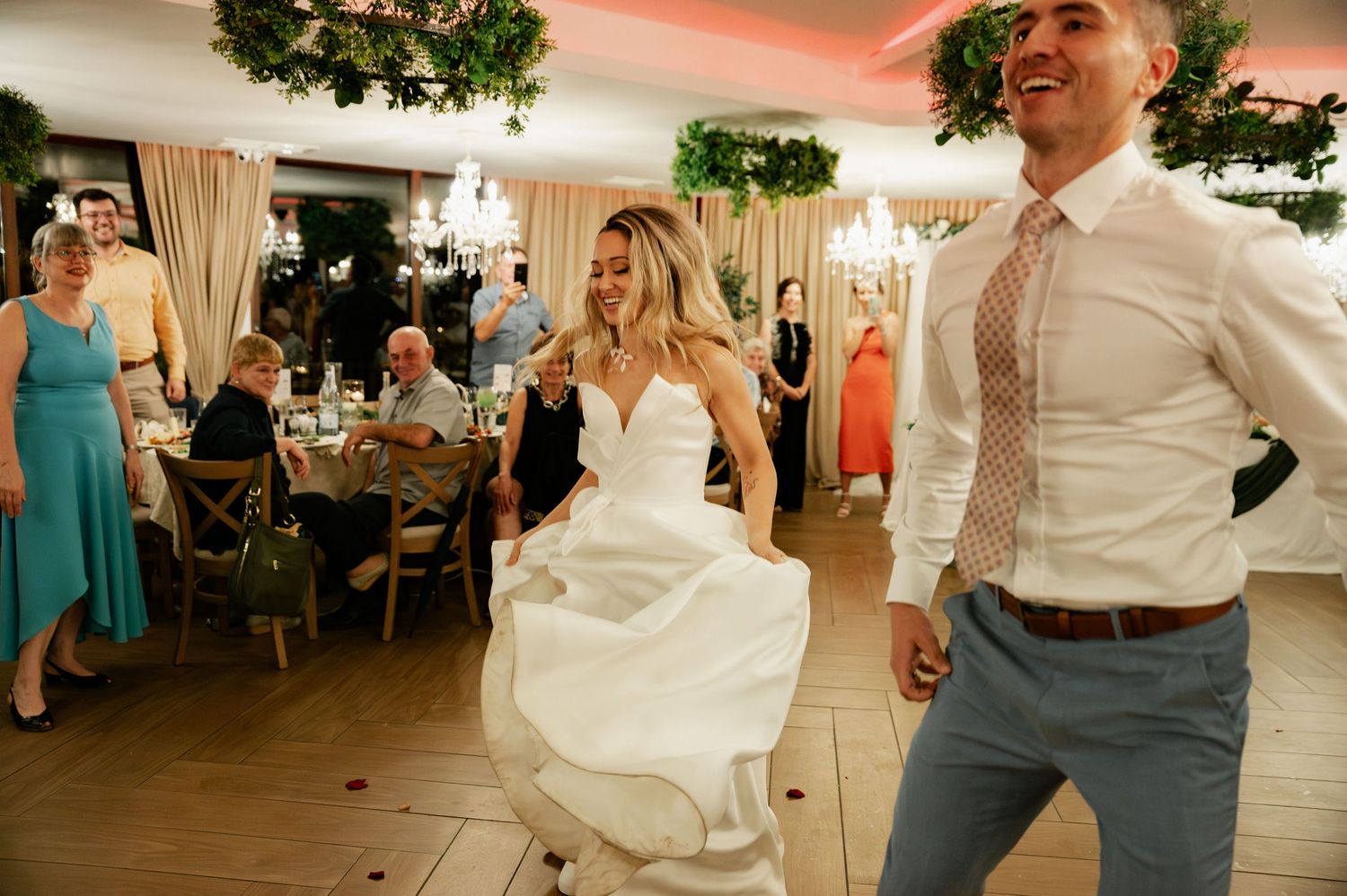 Wedding guests dance and celebrate at an indoor reception venue decorated with hanging greenery and chandeliers.