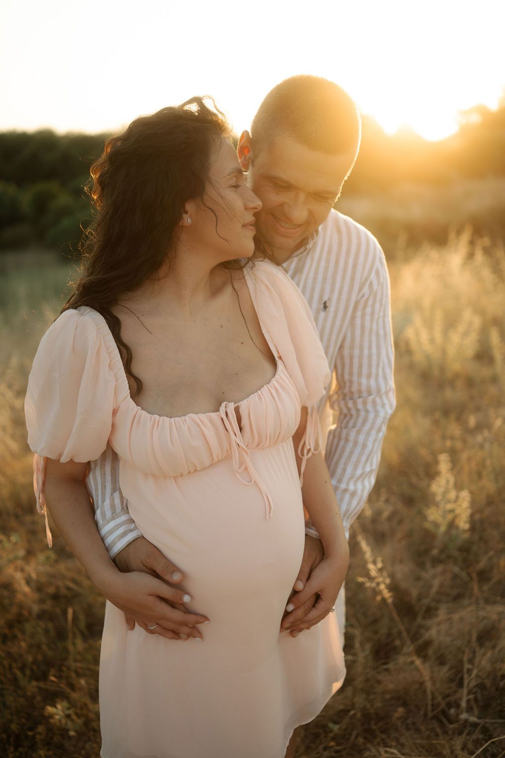 Romantic maternity photoshoot in a golden field at sunset with couple embracing in soft natural light.