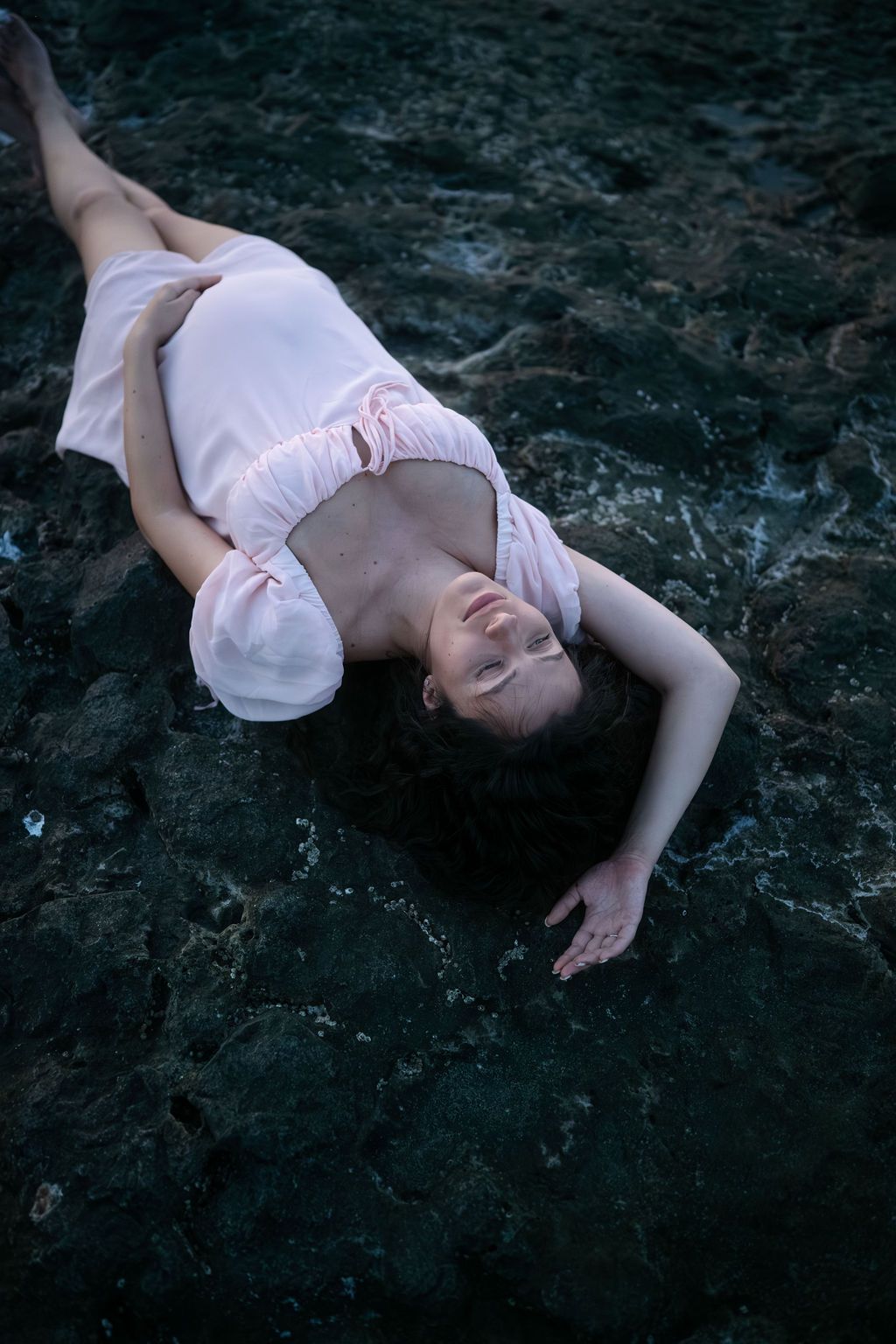Figure in white dress lying on dark rocky surface in artistic dramatic pose.