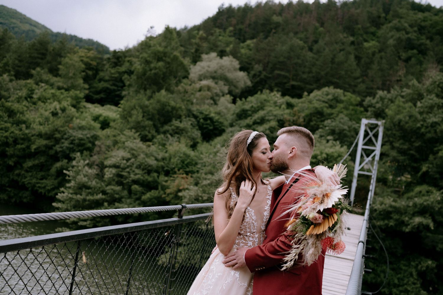 A romantic wedding couple shares a kiss on a suspension bridge surrounded by lush green forest scenery.