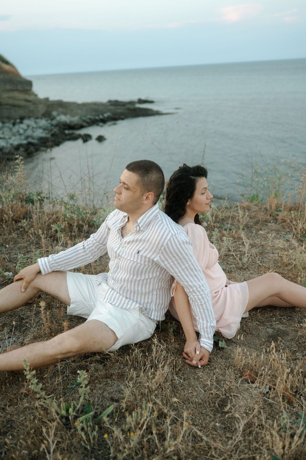 A couple sits back-to-back on a grassy cliff overlooking the ocean during sunset.