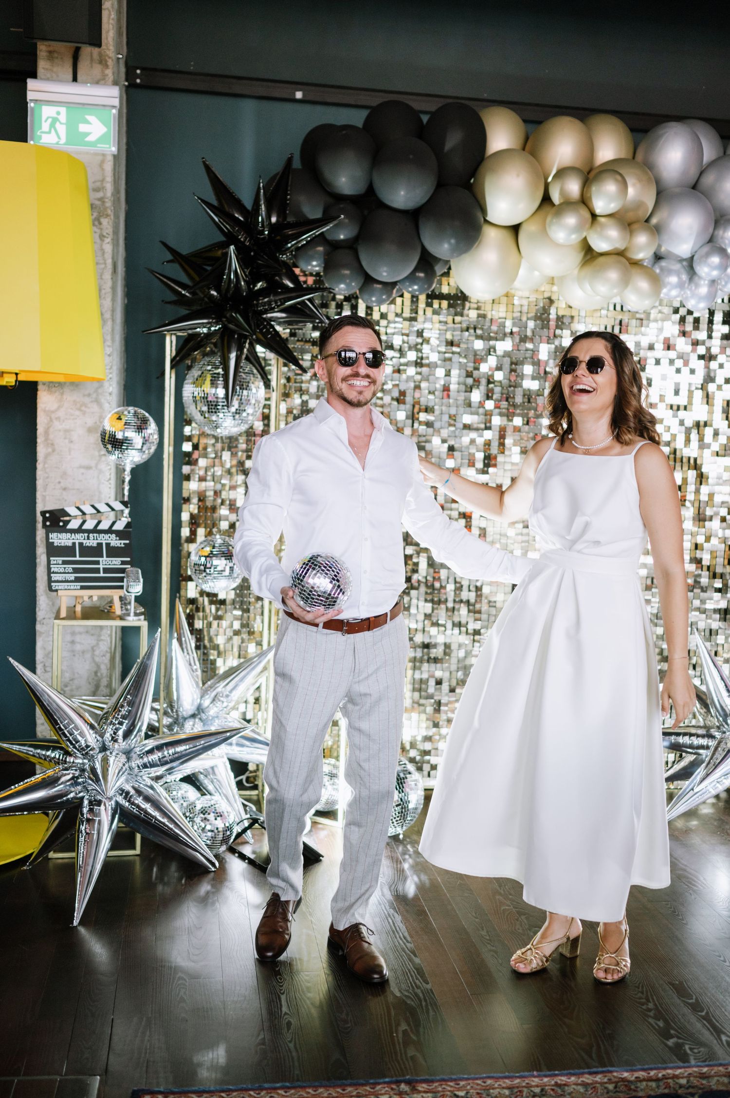 A couple in white outfits and sunglasses poses playfully against a backdrop of silver balloons and decorative stars.