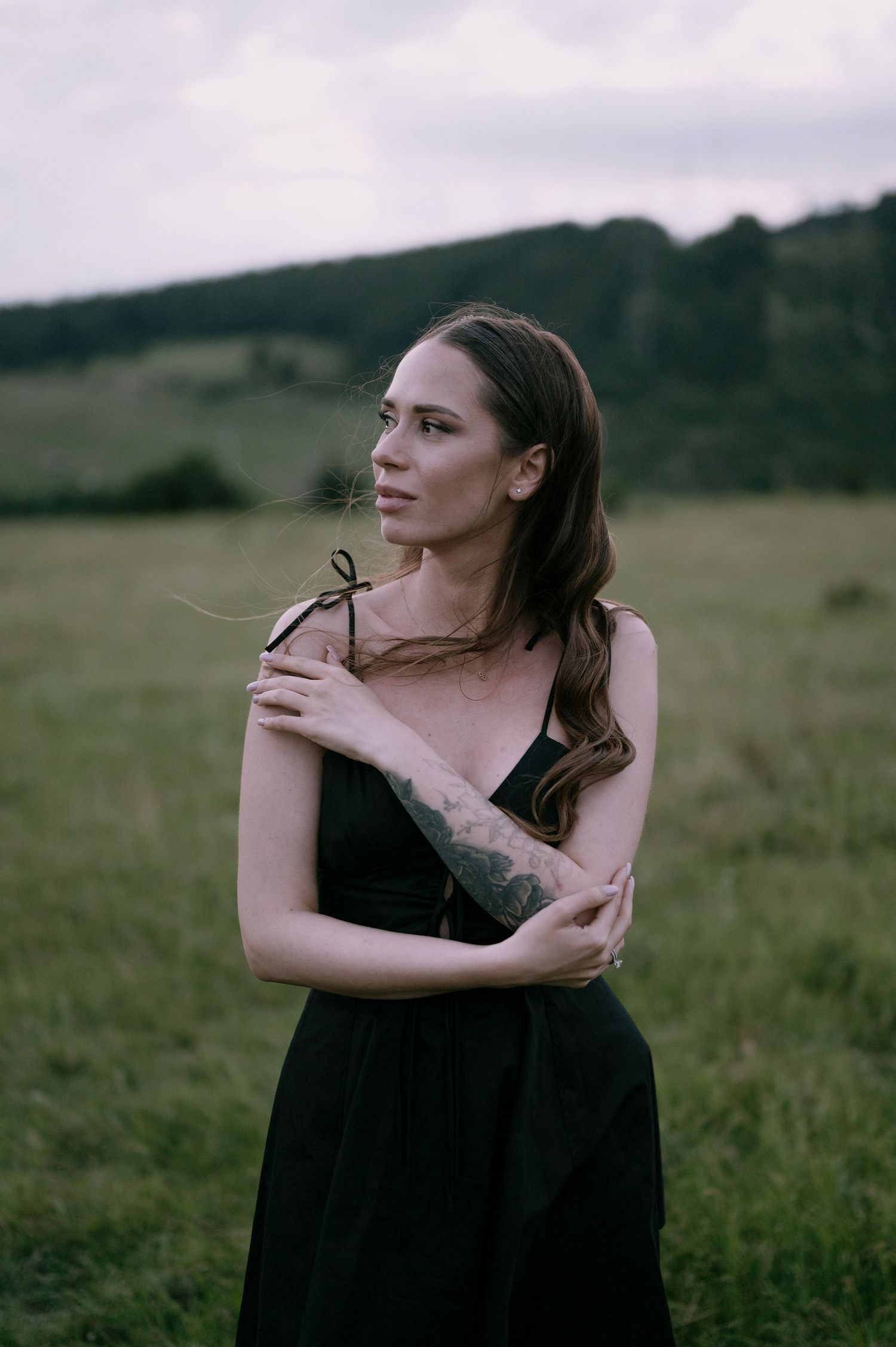 A person in a black dress stands thoughtfully in a grassy field on an overcast day.
