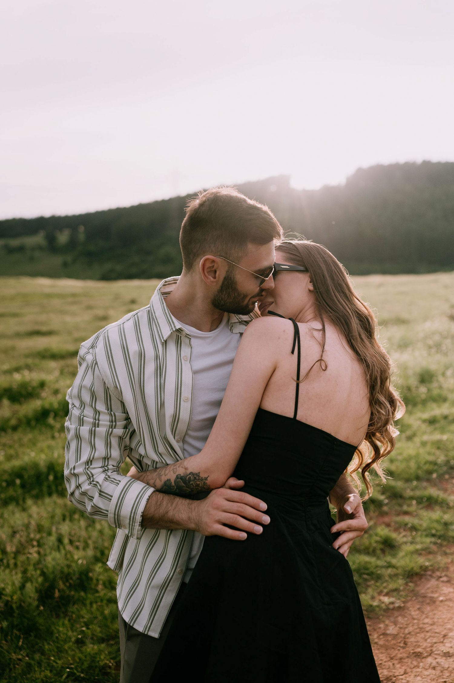 A romantic couple embraces and kisses in a sunlit field during golden hour.