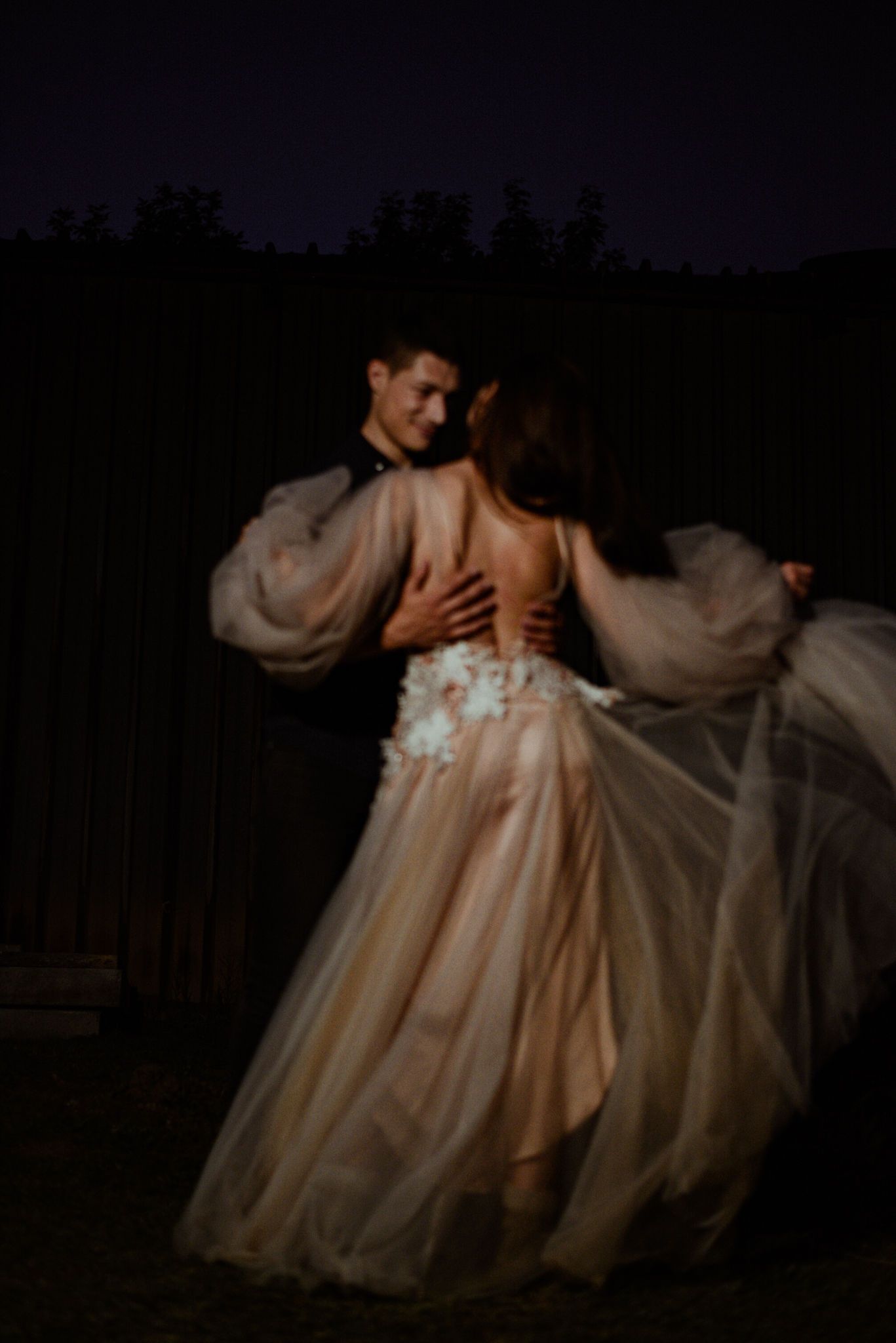 A romantic couple in flowing cream-colored formal attire dance together under dark evening skies.