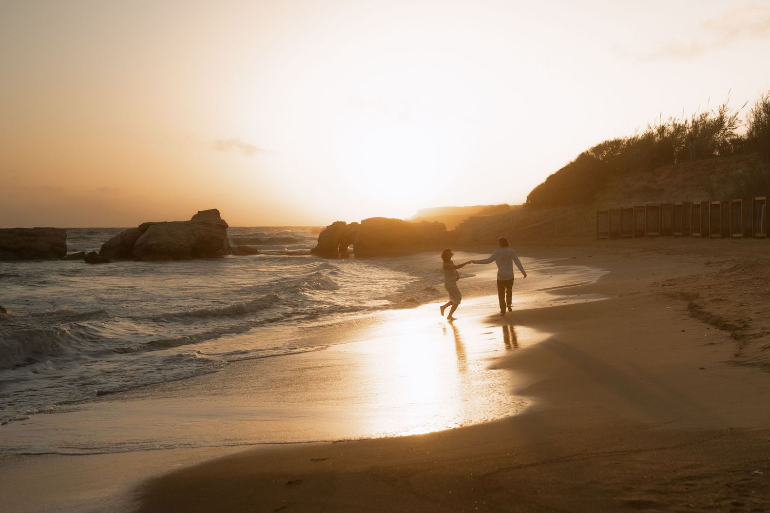 Two silhouetted figures walk along a sandy beach at golden sunset with ocean waves and rocky cliffs in background.